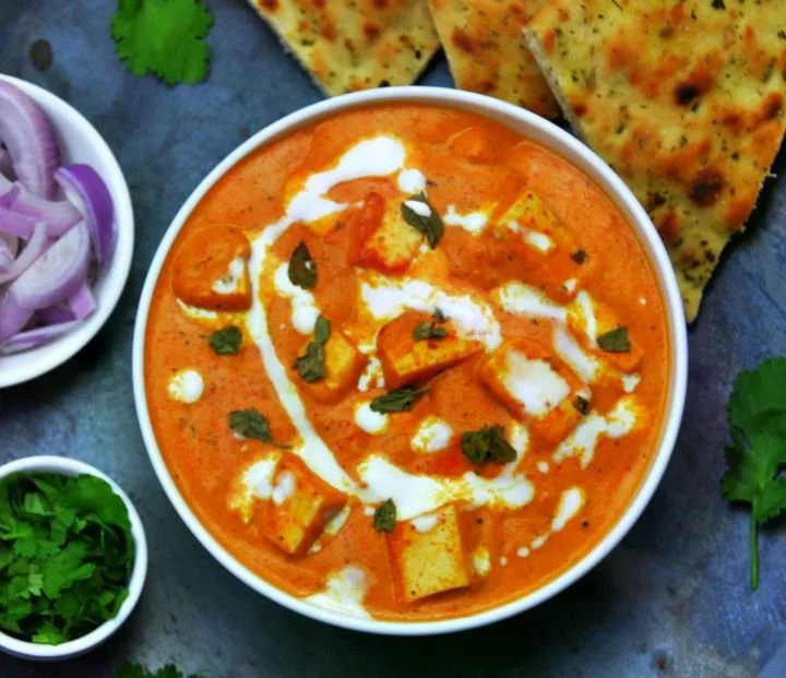 A bowl of Indian paneer tikka masala with white cream drizzle garnished with cilantro, served with naan bread, alongside small bowls of sliced red onions and cilantro leaves.