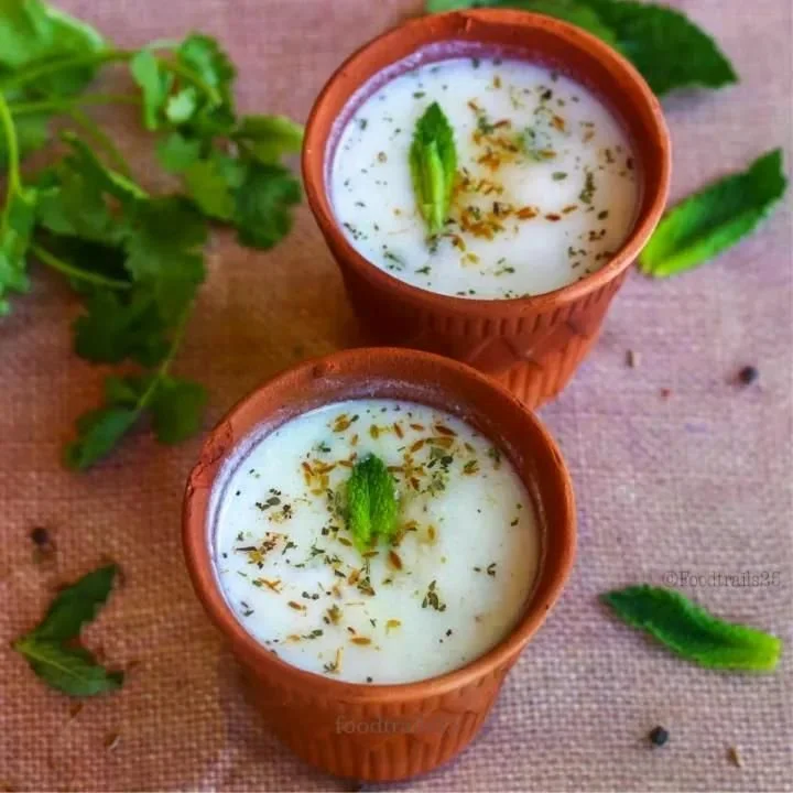 Two clay cups filled with a creamy white Indian dessert garnished with green garnish and dry herbs, on a rustic cloth surface with scattered green leaves around.