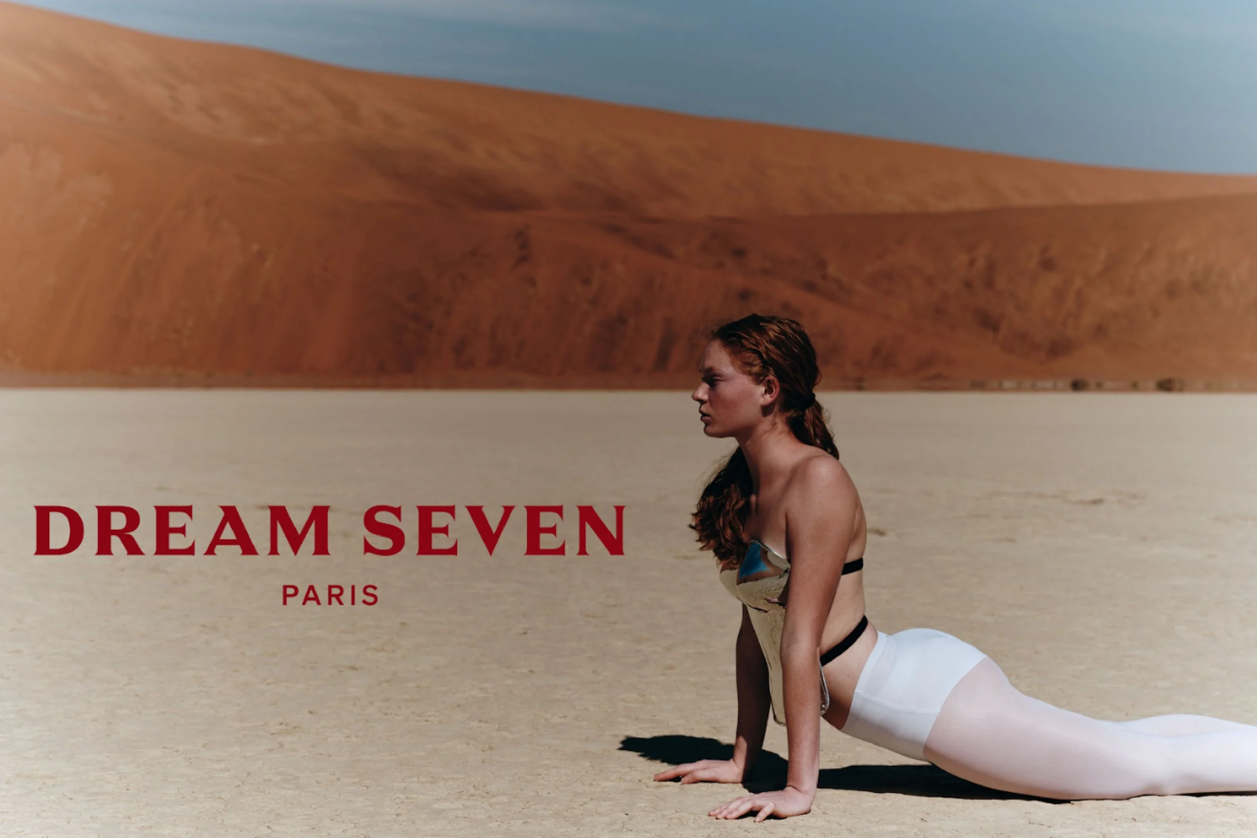 A woman in a yoga pose on a sandy desert with orange dunes and a blue sky in the background. The text reads "DREAM SEVEN PARIS."