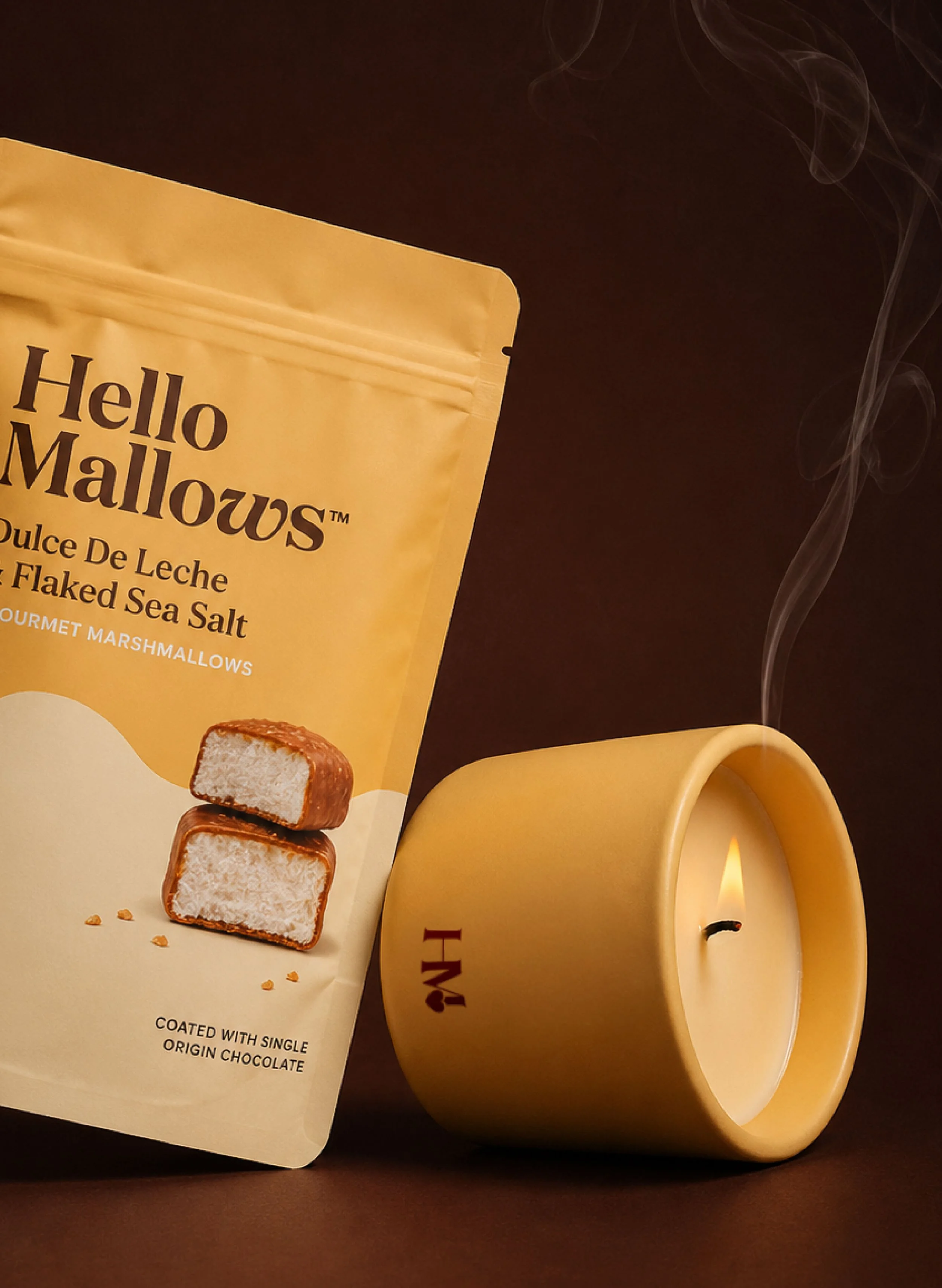 A yellow package of Hello Mallow's toasted coconut flavored marshmallows, with two marshmallows stacked in front of the package and a lit candle in a yellow holder to the right.