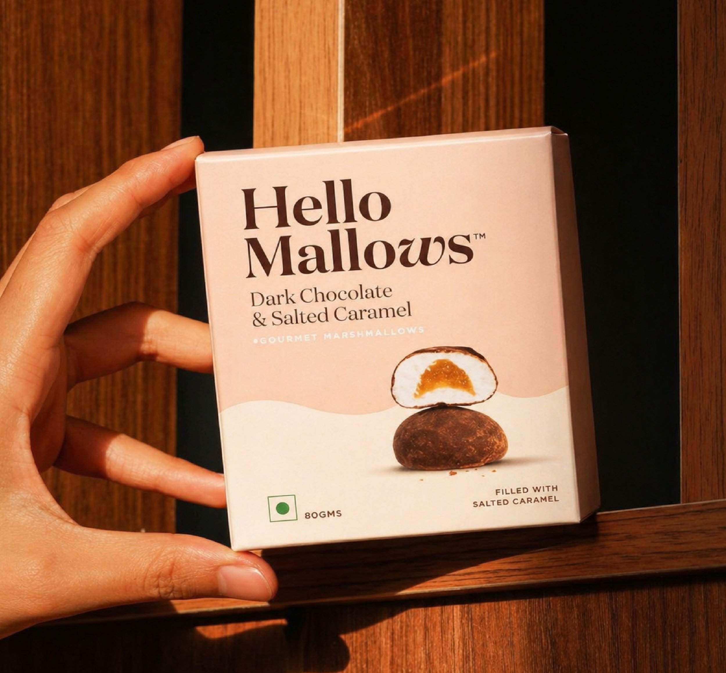 A hand holding a box of Hello Mallows dark chocolate and salted caramel flavored marshmallows against a wooden background.