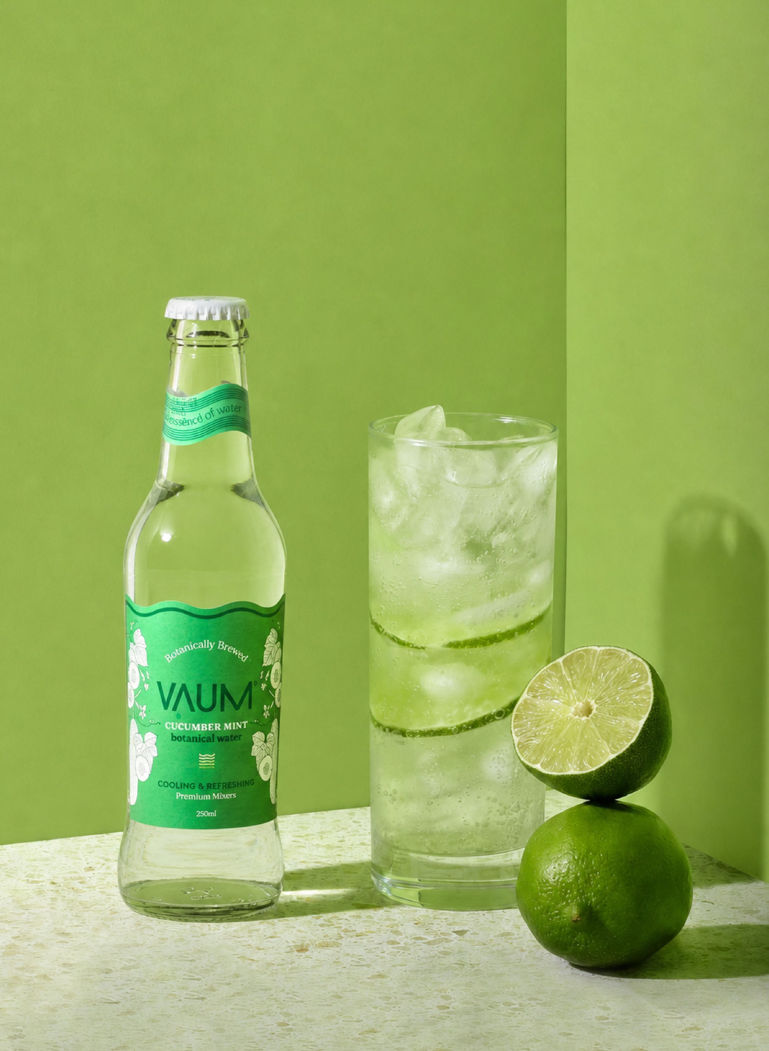 A bottle of cucumber mint flavored botanical water, a glass filled with the same beverage and ice, a halved lime, and a whole lime, all set against a bright green background.
