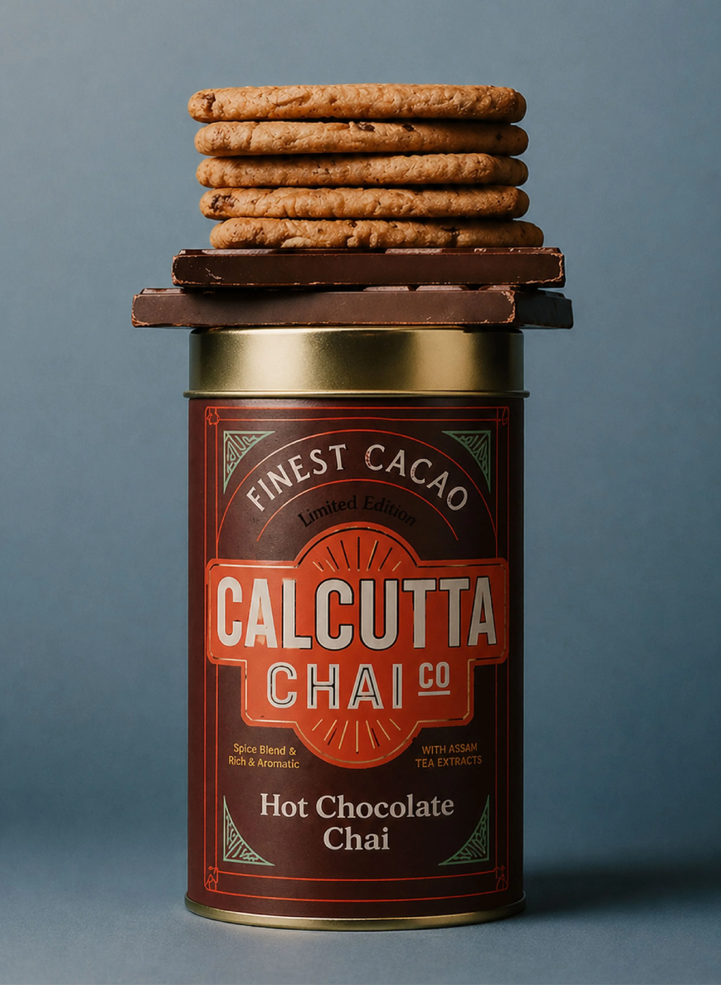 A stack of six cookies and two bars of dark chocolate on top of a tin container with a label for Calcutta Chai. The label mentions hot chocolate chai with Assam tea extracts and describes it as a spice blend, rich and aromatic.