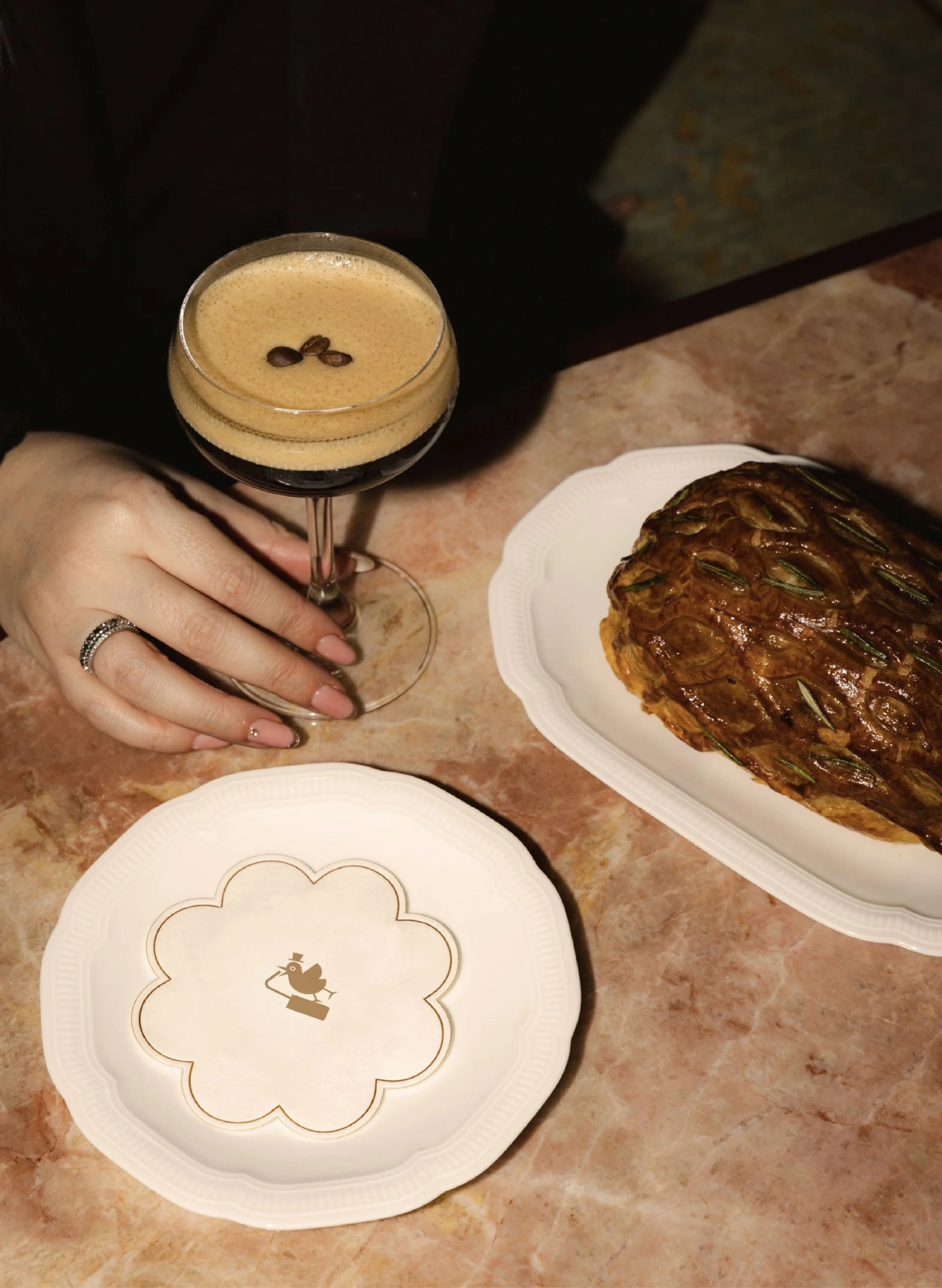 A person holding a glass of espresso with coffee beans on top, next to a pineapple upside-down cake on a white platter, and a decorative paper plate with a bird illustration.