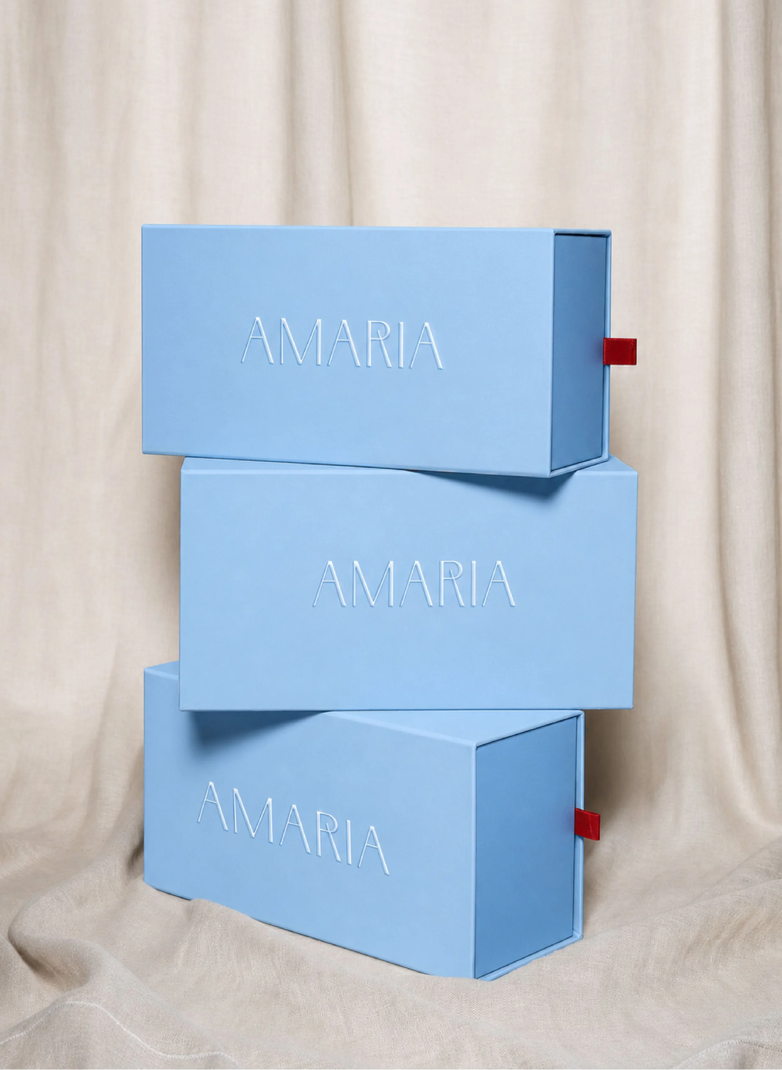 Stacked blue boxes with the word 'AMARIA' embossed on each, set against a beige curtain backdrop.
