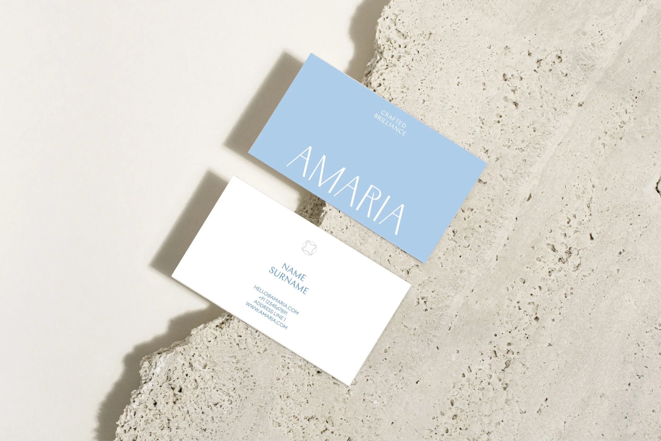 Business cards on textured concrete surface. One card is blue with white text that reads 'CRAFTED BRILLIANCE' and 'AMARIA'. The other card is white with blue text displaying placeholder contact information.
