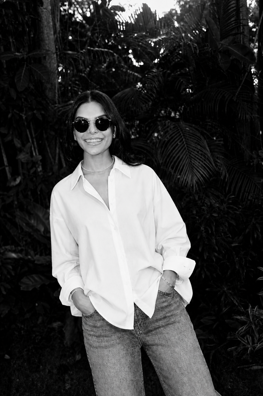 A woman smiling outdoors, wearing sunglasses, a white shirt, and jeans, with her hands in her pockets, in front of lush foliage.