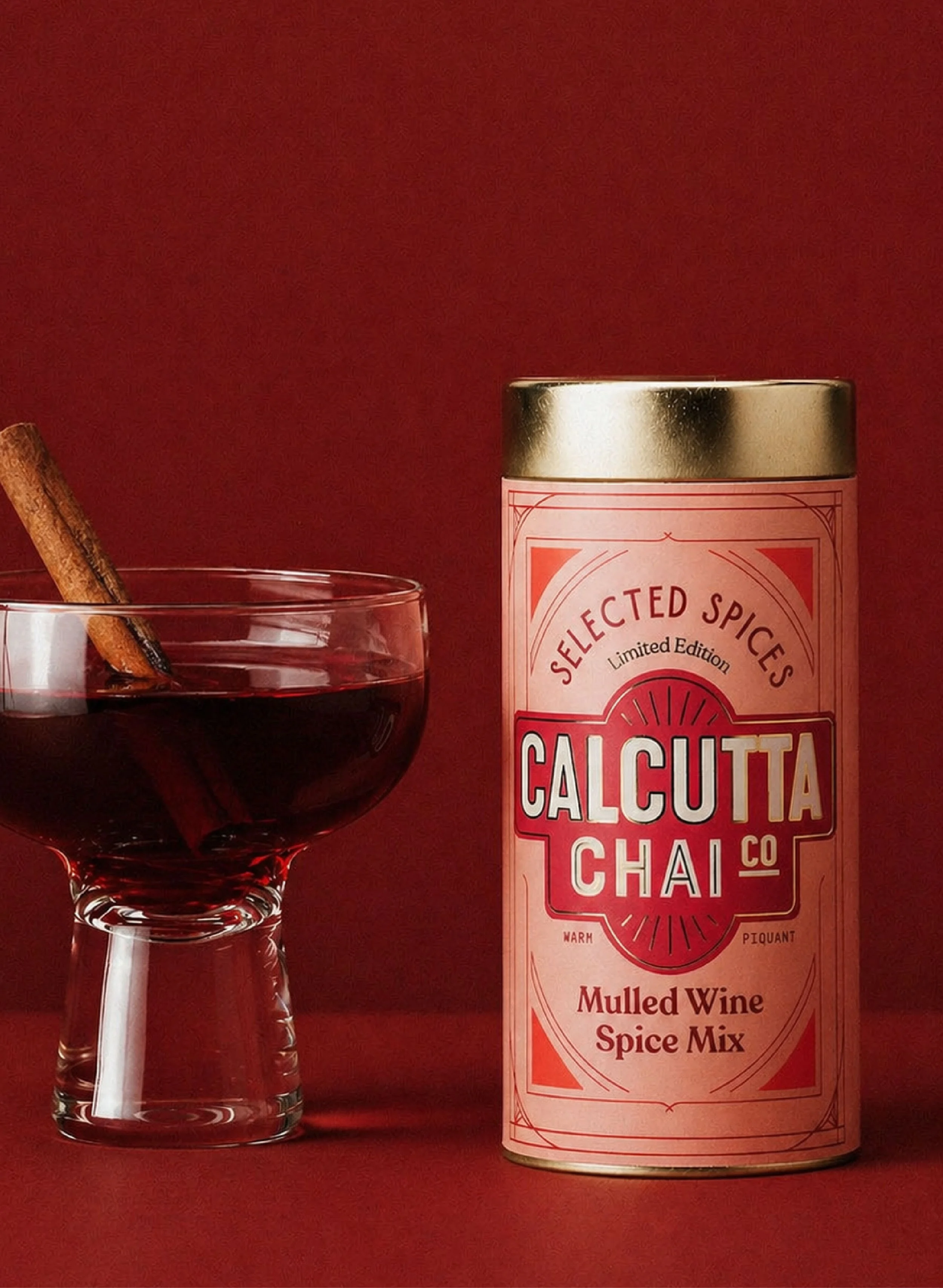 A glass of red mulled wine with a cinnamon stick on the rim, next to a can of Calcutta Chai mulled wine spice mix, all set against a red background.