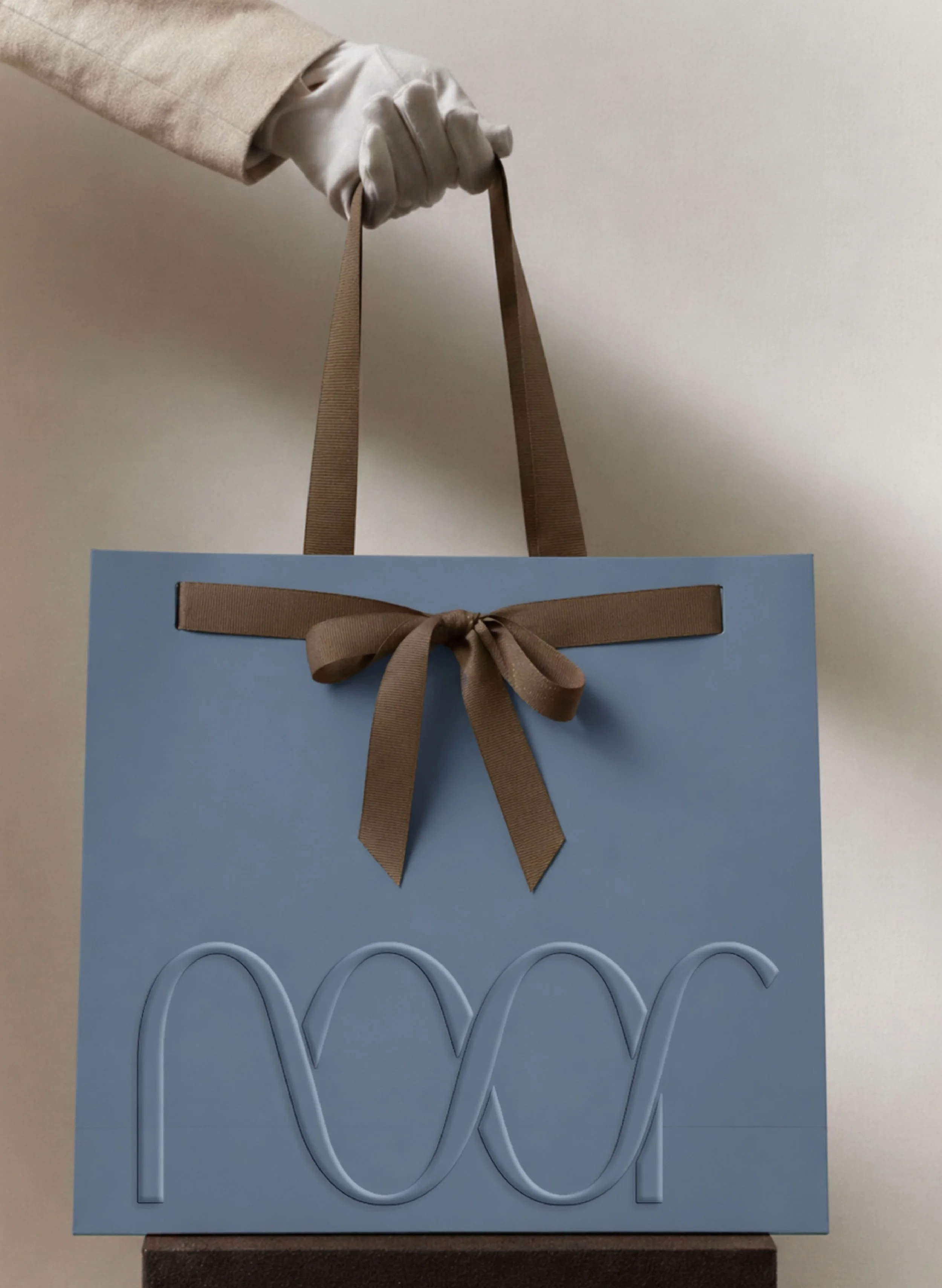 A light blue gift bag with a brown ribbon bow on the front, containing three white heart-shaped lollipops with curved sticks, set against a light-colored background.