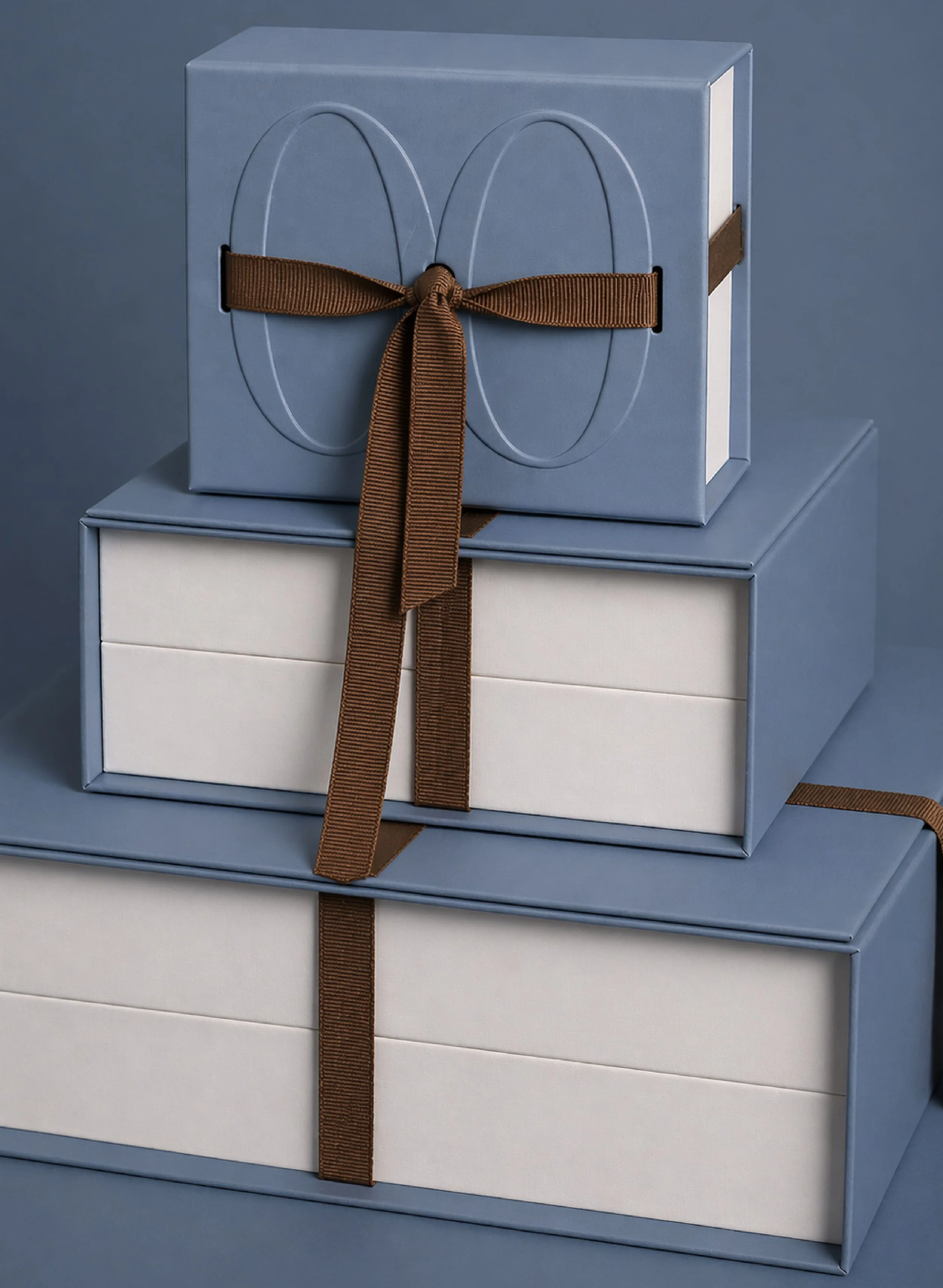 A stack of three gift boxes, each with a ribbon. The top box is blue with a brown ribbon, the middle box is blue with a brown ribbon, and the bottom box is white with a blue ribbon.