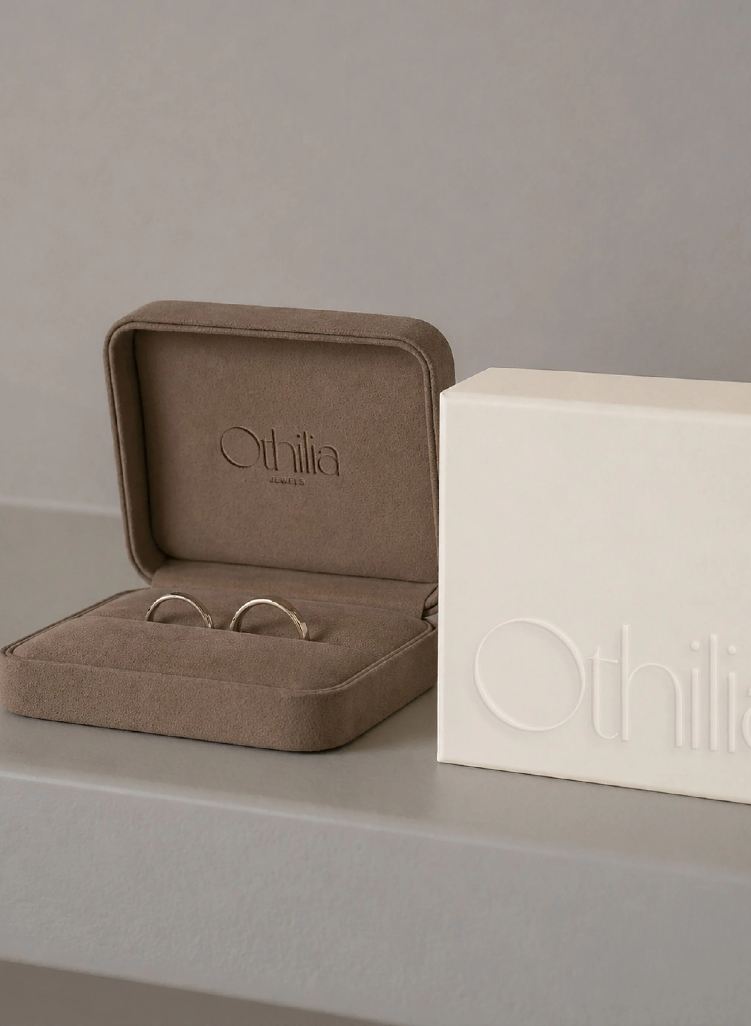 A beige jewelry box with the brand name 'Othilia Jewels' embossed inside the lid, open to reveal two silver rings, next to a white box with faint embossed text.