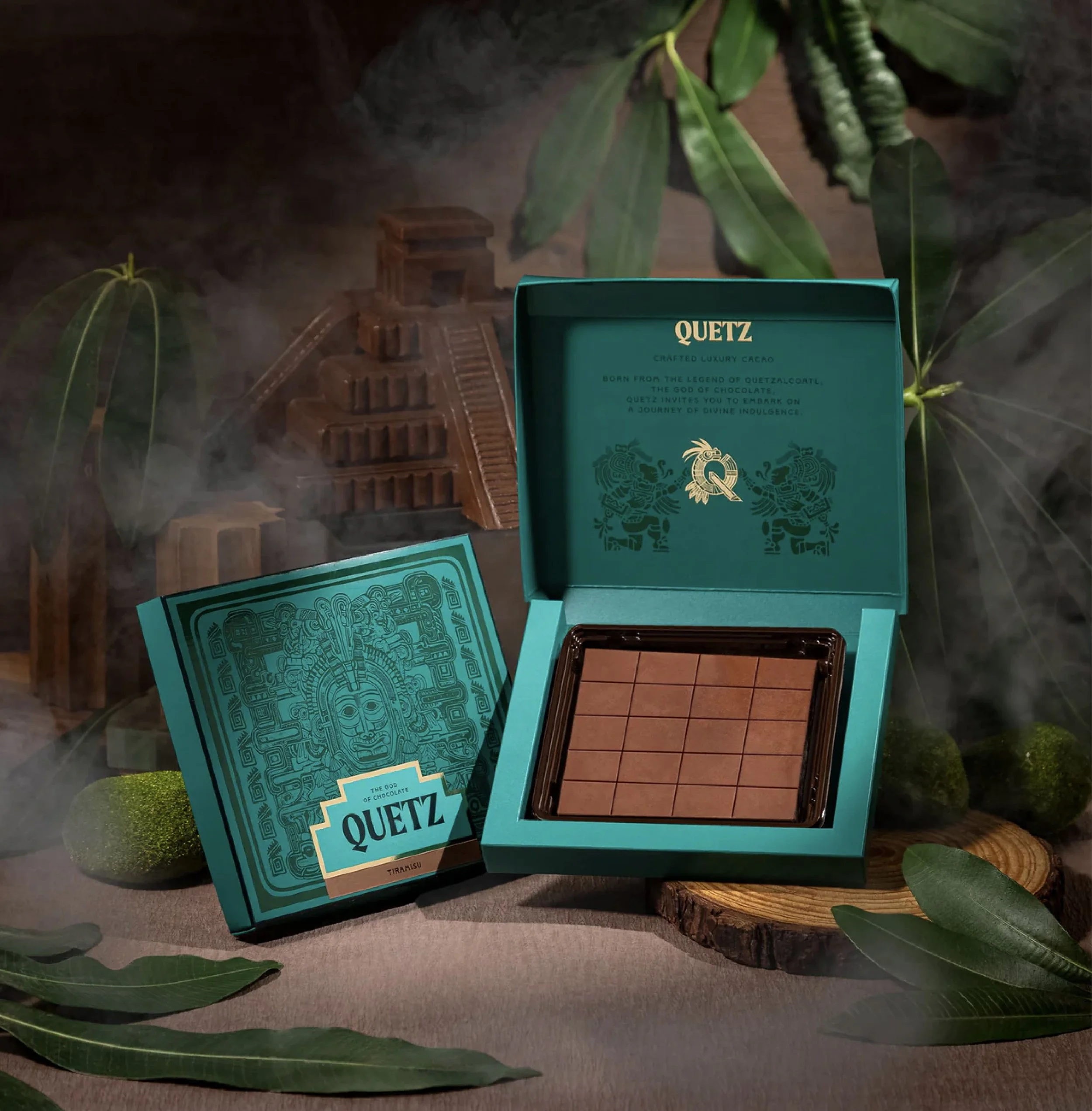 Open chocolate box labeled Quetz with a picture of a serpent, and a matching blue panel behind it. The box contains smaller chocolate pieces in a grid pattern. There are large green leaves and a wooden log beneath the box, with a background featuring