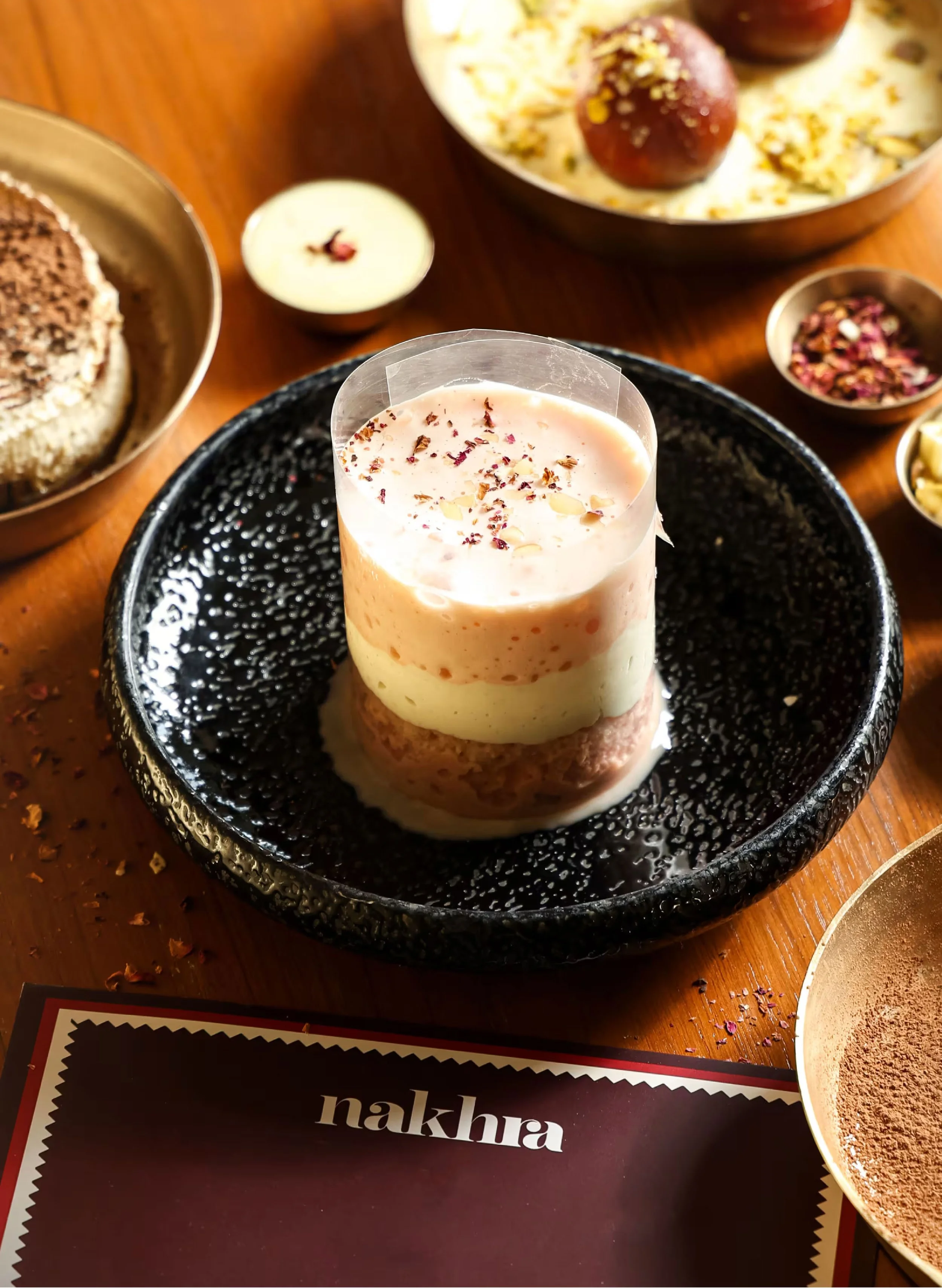 Close-up of a layered dessert topped with rose petals and dry flower petals, in a glass on a black plate with a textured surface, surrounded by bowls of sweets and small containers with toppings on a wooden table.