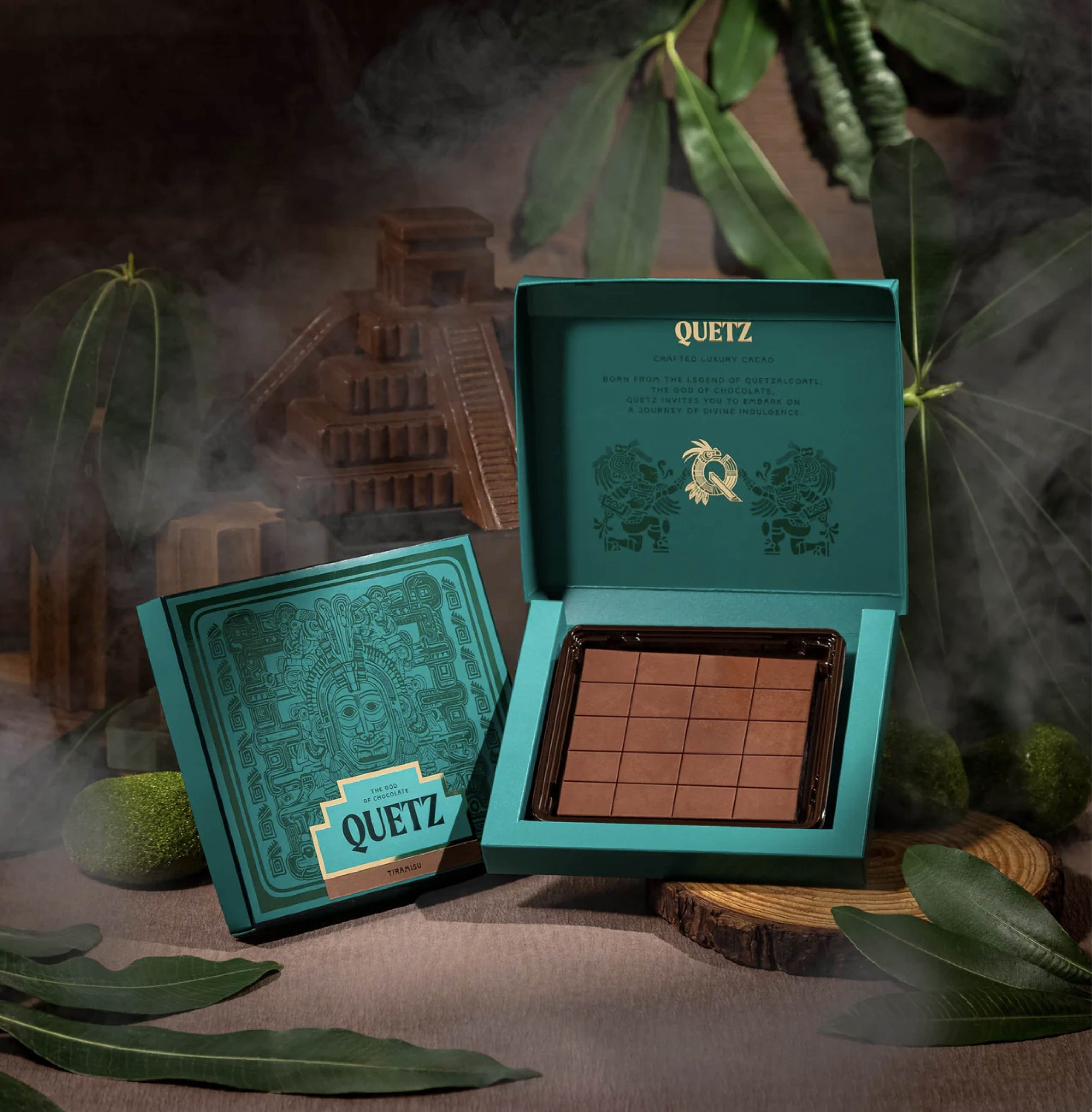 Open teal box with chocolate brownies on a wooden base, surrounded by green leaves, and a teal card with gold and white text that reads "QUETZ" and describes crafted luxury cacao.