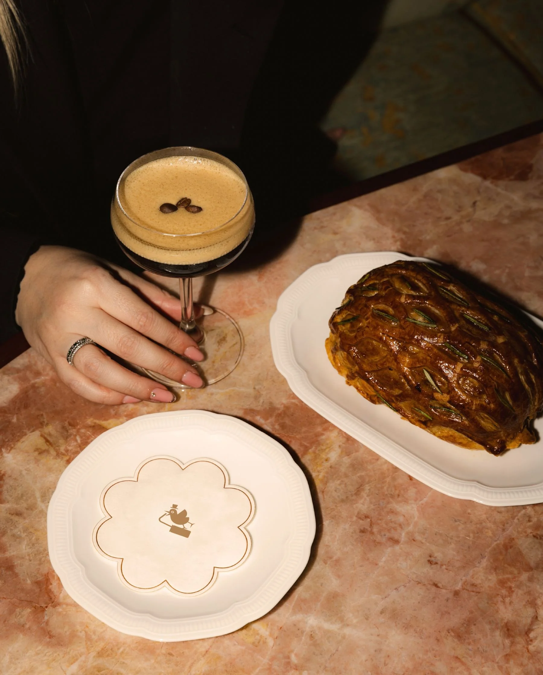 A person holding a coffee drink with coffee beans on top, next to a pineapple on a white plate, set on a marble table.