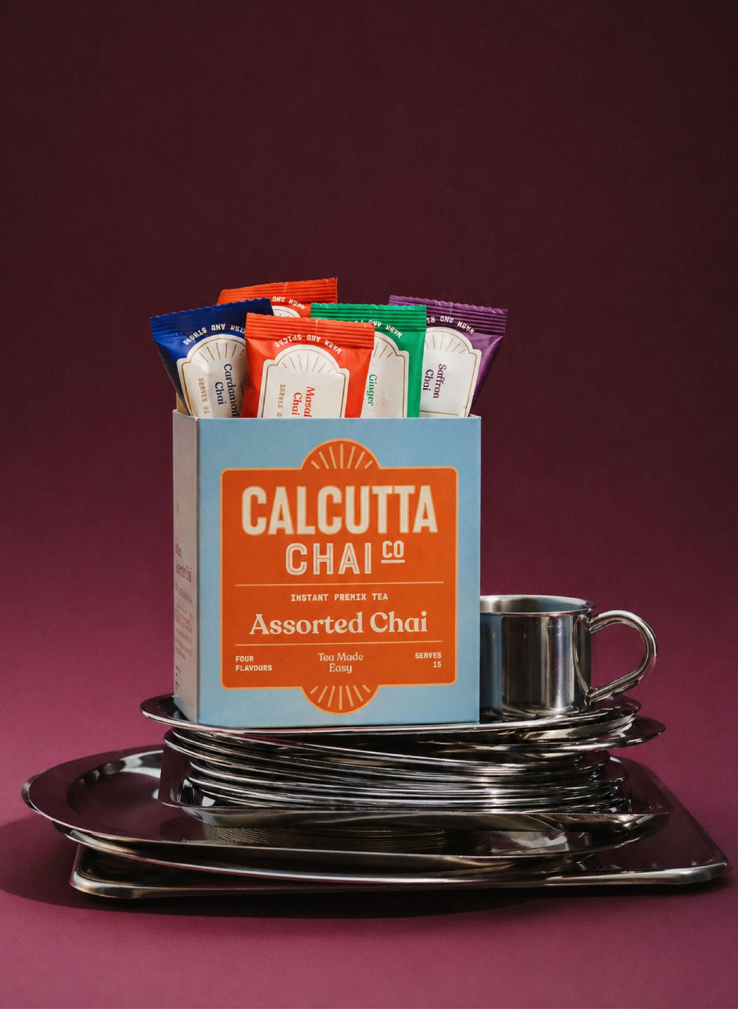 Box of assorted chai tea packets and a stainless steel mug on a reflective tray against a dark pink background.