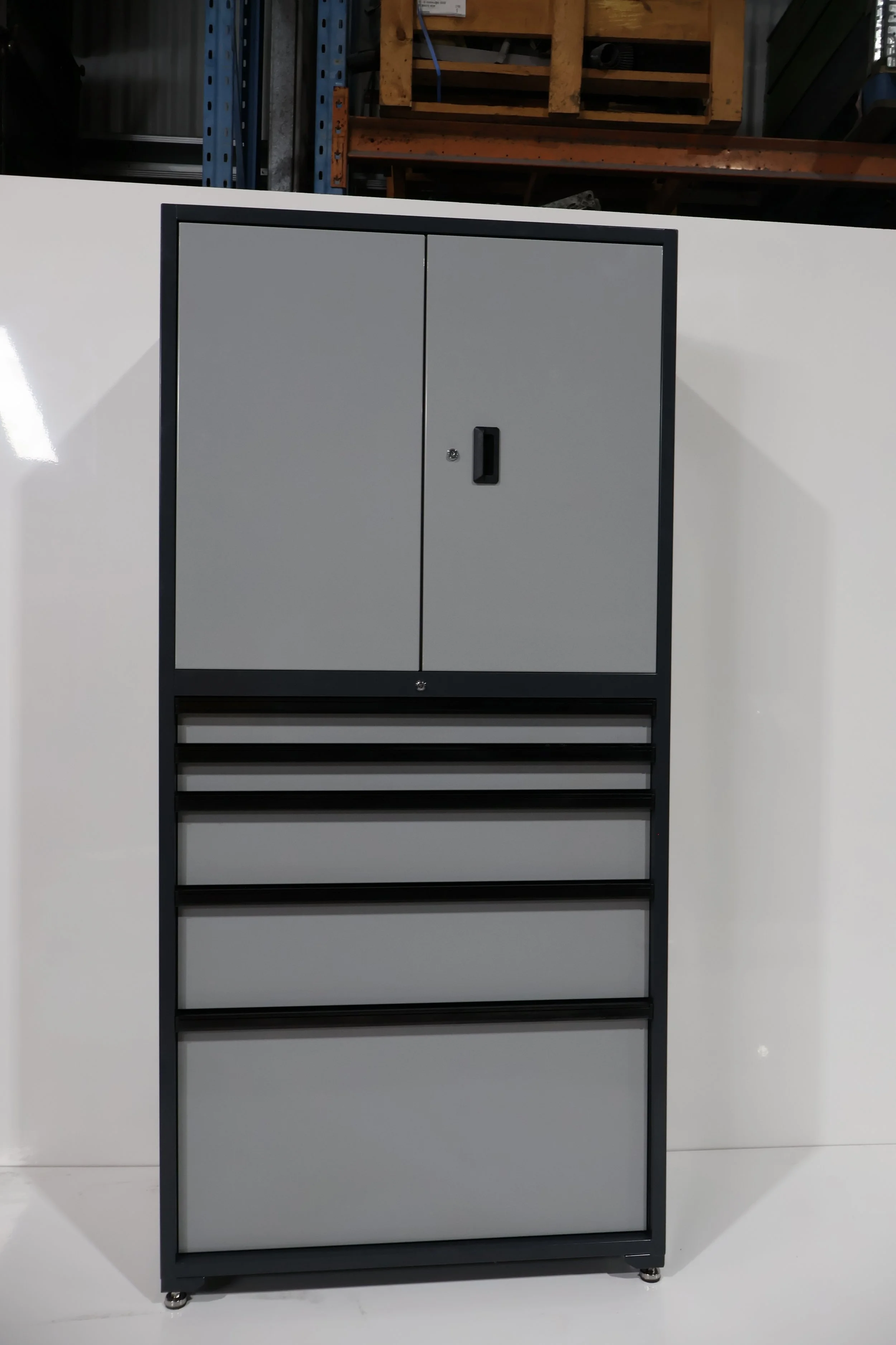 A gray storage cabinet with two doors on top, black trim, and four drawers at the bottom. It is placed against a white background with a warehouse or industrial setting visible in the background.