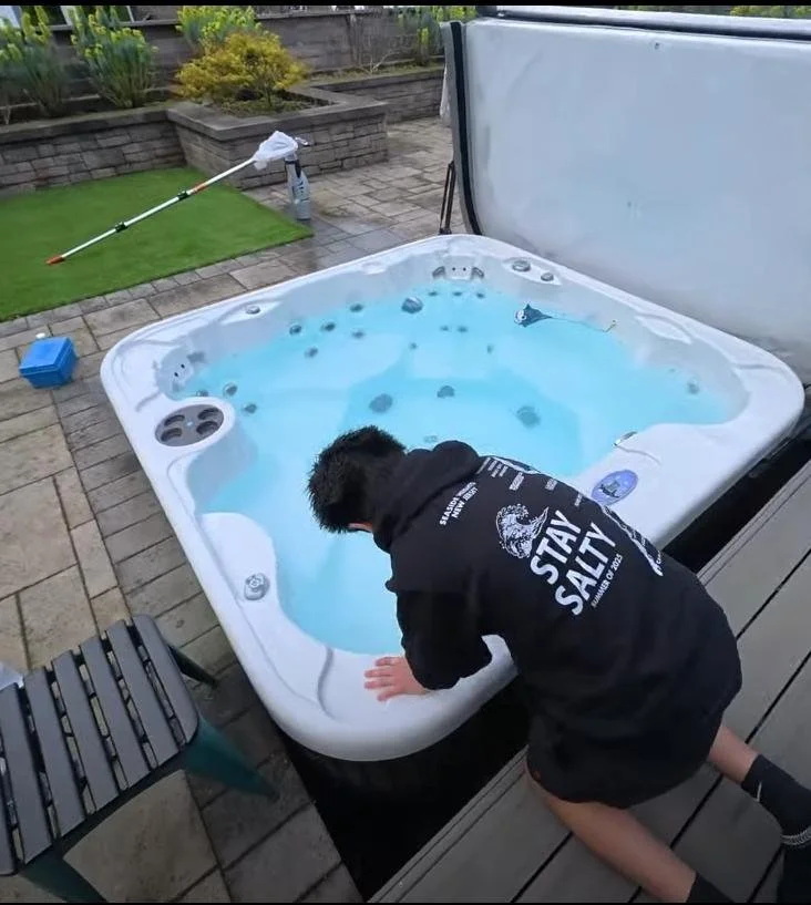 A Pool Technician in a black hoodie with the words 'Stay Salty' leaning over the edge of a hot tub filled with water, outdoors on a patio.