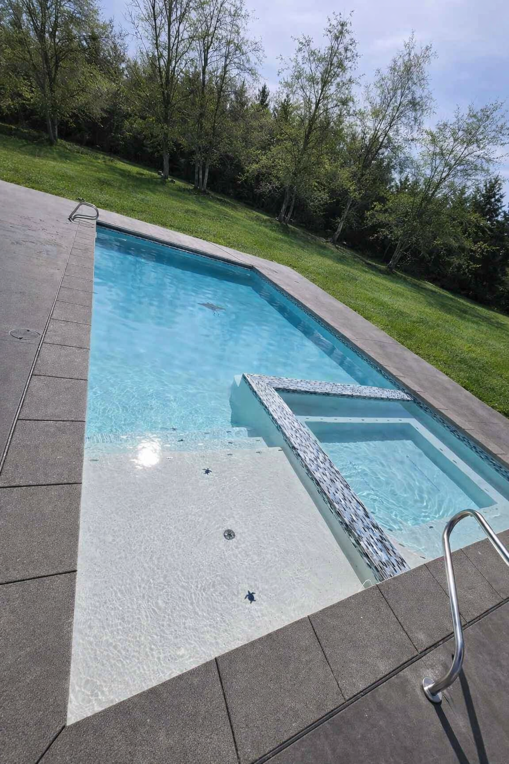 A backyard swimming pool with a built-in hot tub, surrounded by concrete and a grassy area with trees in the background, under a partly cloudy sky.