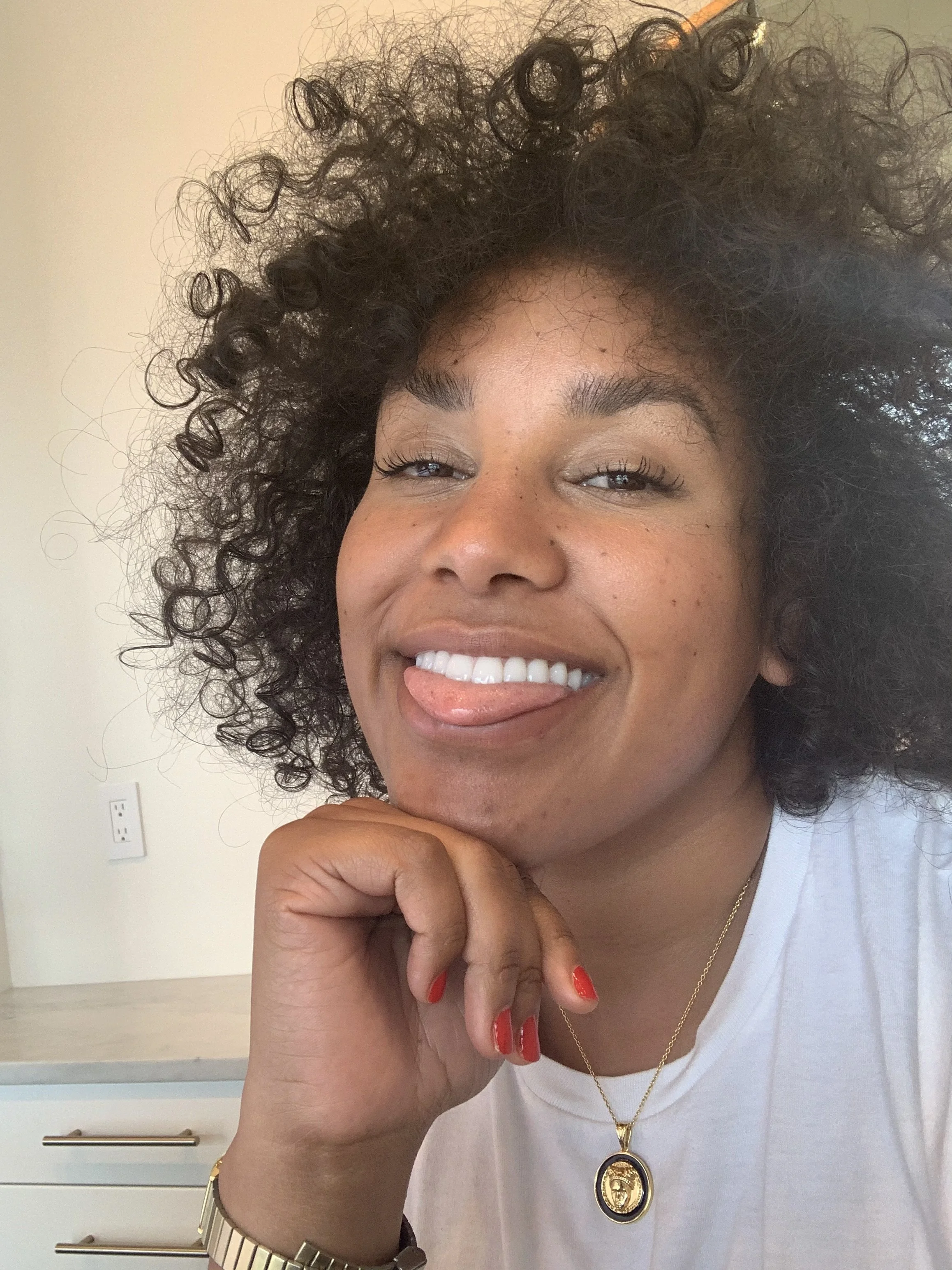 A woman with curly hair sticking her tongue out and smiling, resting her chin on her hand, wearing a white shirt, gold necklace with a pendant, and a gold watch, indoors with a plain wall and a white electrical outlet in the background.