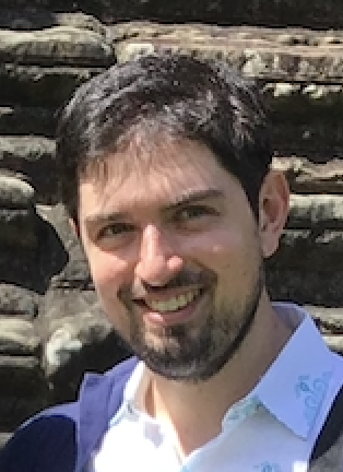 A smiling man with dark hair and a beard outdoors, wearing a white shirt and blue jacket, with a stone staircase in the background.