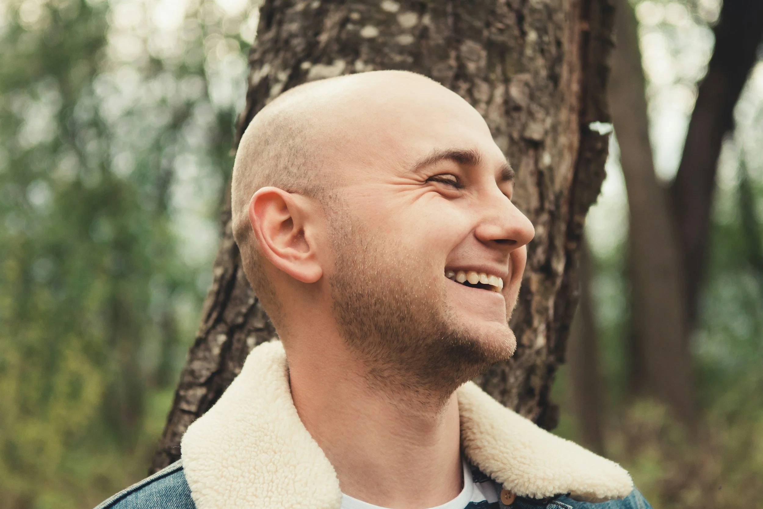 A smiling man with a shaved head and beard standing outdoors near a tree, wearing a jacket with a shearling collar, in a forested area.