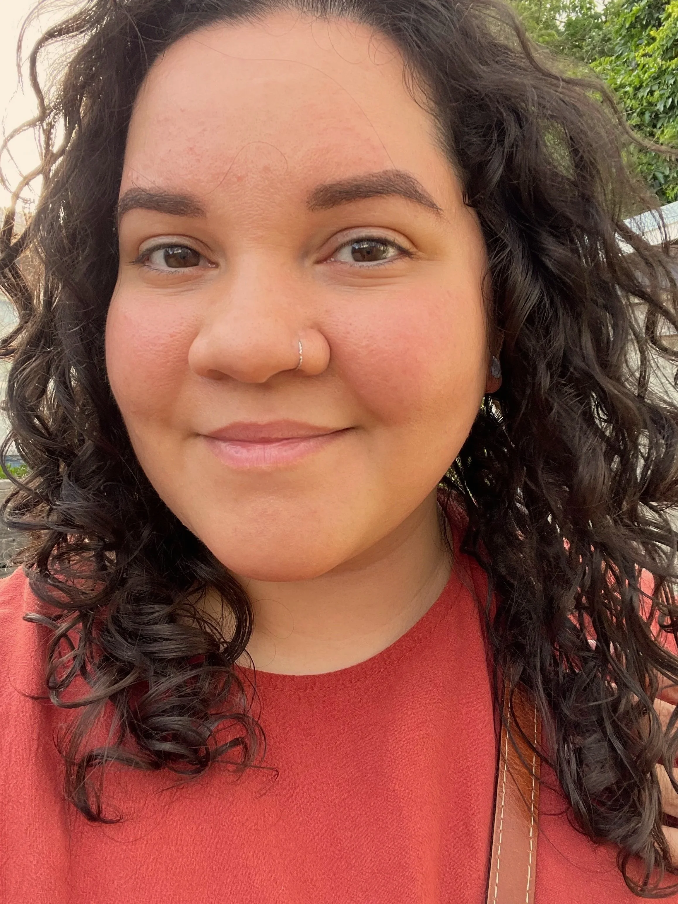 Close-up of a woman with curly dark hair, a nose piercing, wearing a red top, outdoors with green foliage in the background.