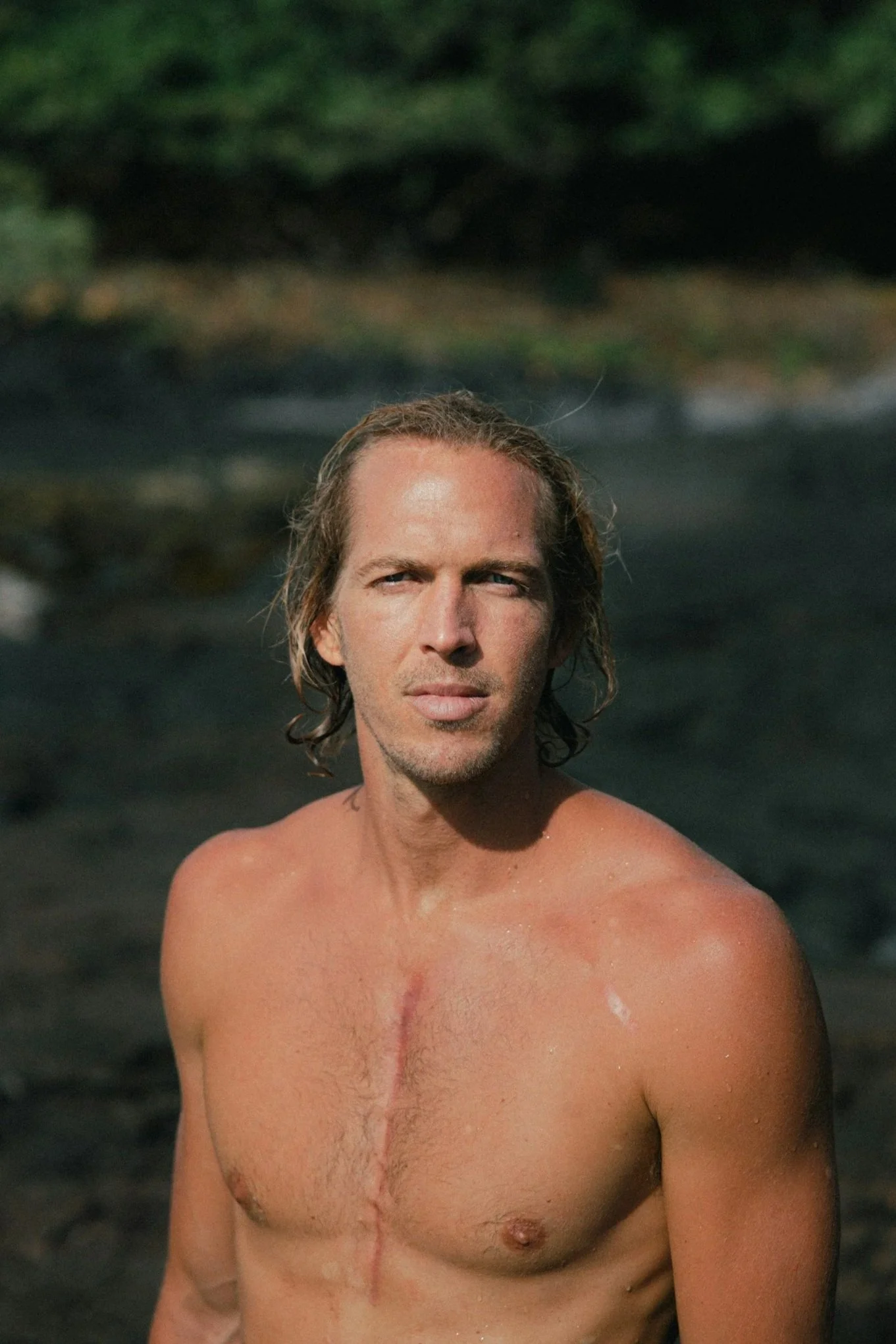 A shirtless man with long hair standing outdoors with a chest scar, possibly near water, with a blurred background of greenery.