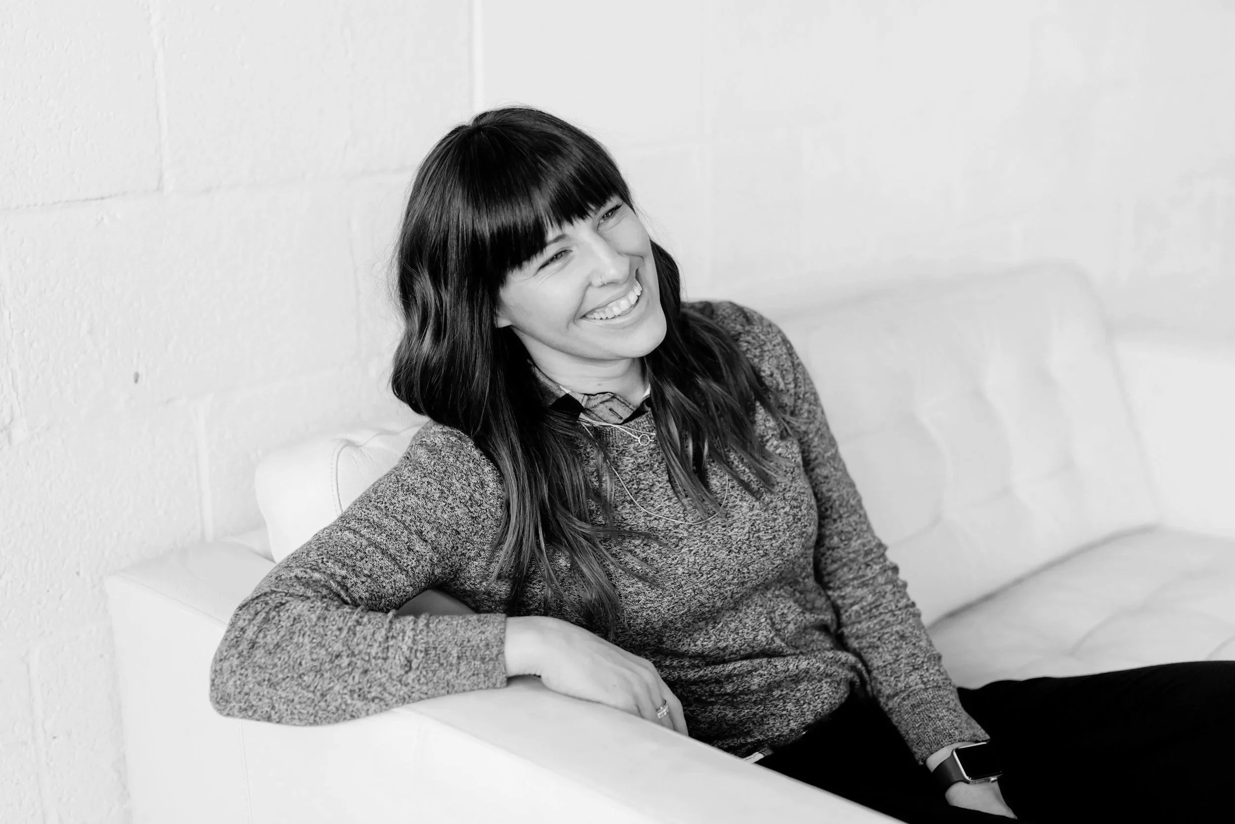 Black and white photo of a woman with long dark hair and bangs sitting on a white bench against a white brick wall, smiling and looking to her right.