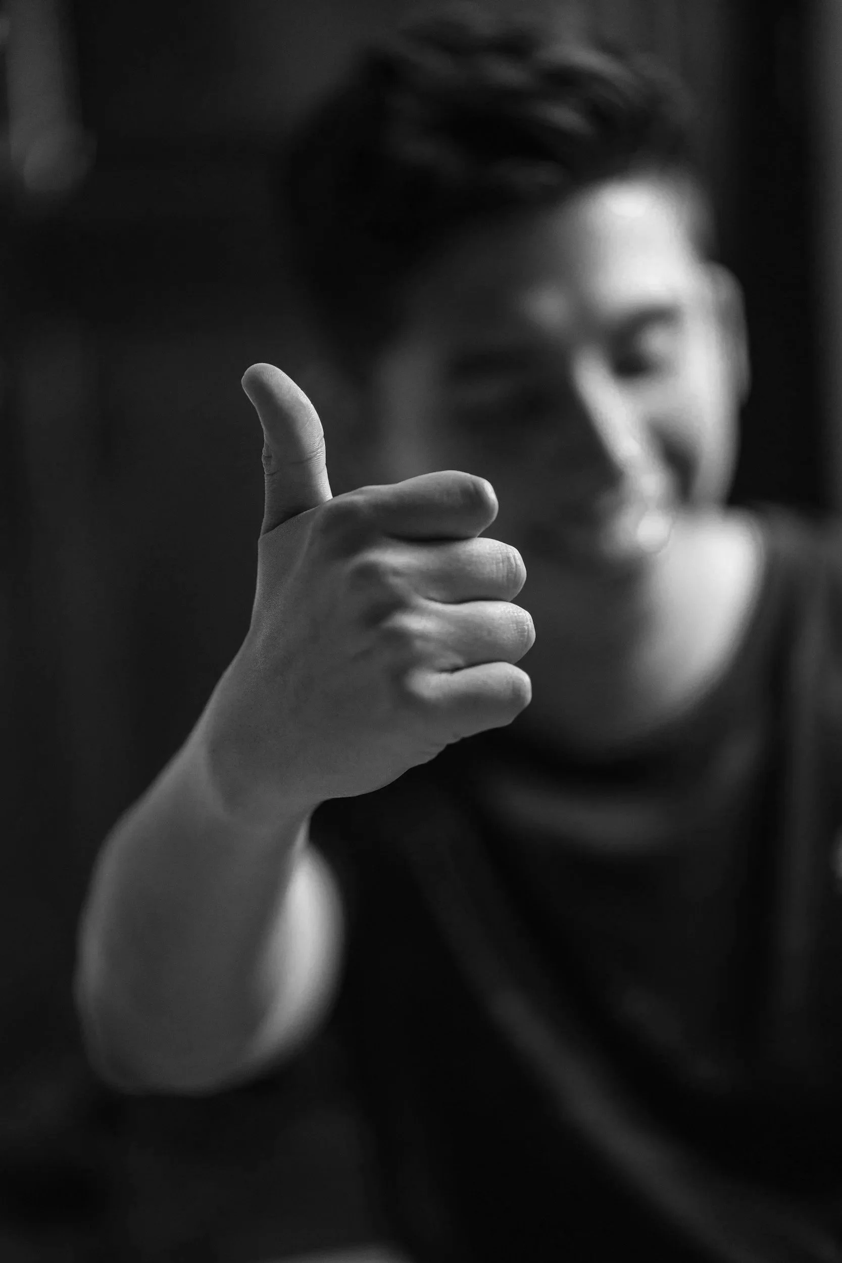 A black and white photo of a person smiling, with their face slightly out of focus, and holding their thumb up in a gesture of approval.