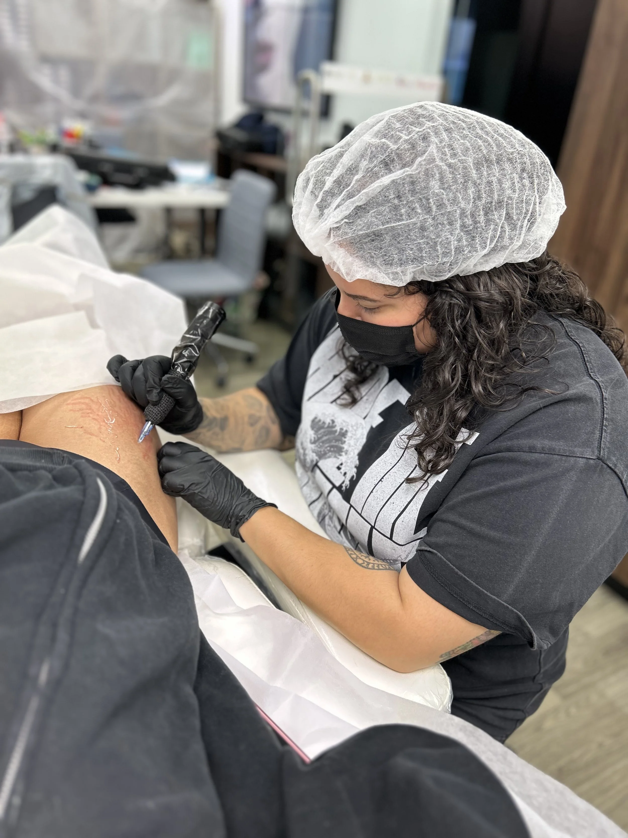 A person wearing a hairnet, black face mask, and black gloves is tattooing a person's arm in a clinical setting.