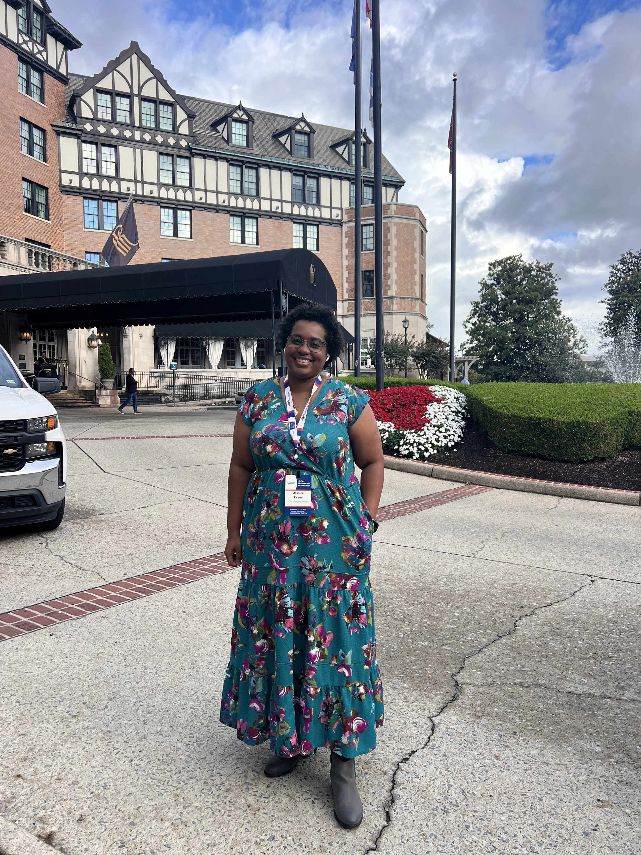 Jessica Clérc Evans standing outside the Hotel Roanoke during the 2024 LAP Conference, wearing a teal dress and conference badge, with landscaped grounds and flags in the background.