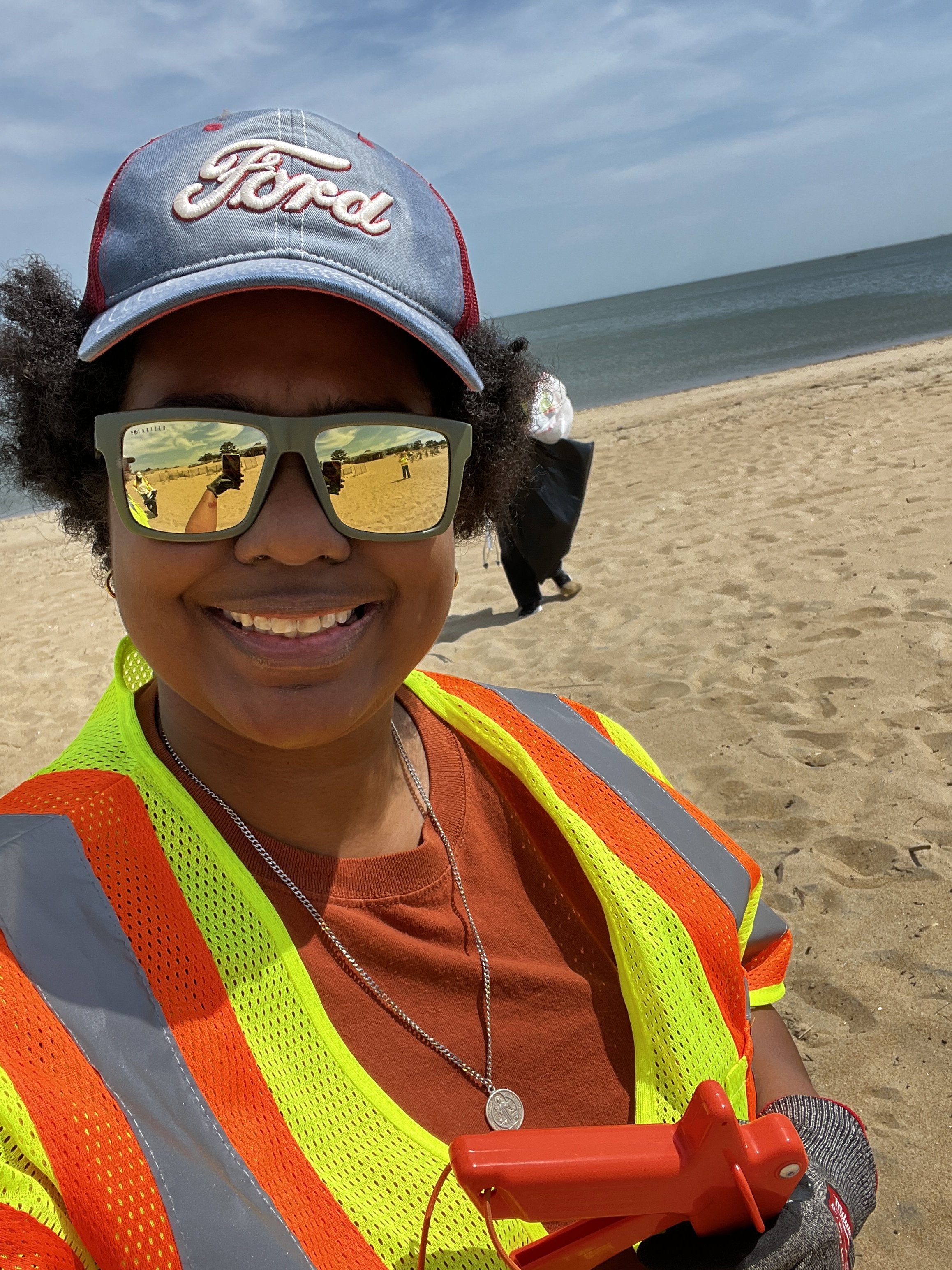 A person wearing a Ford baseball cap, sunglasses, a neon green safety vest, and an orange shirt at the beach, holding a red device with a handle and a cord, with the ocean and sandy beach in the background.
