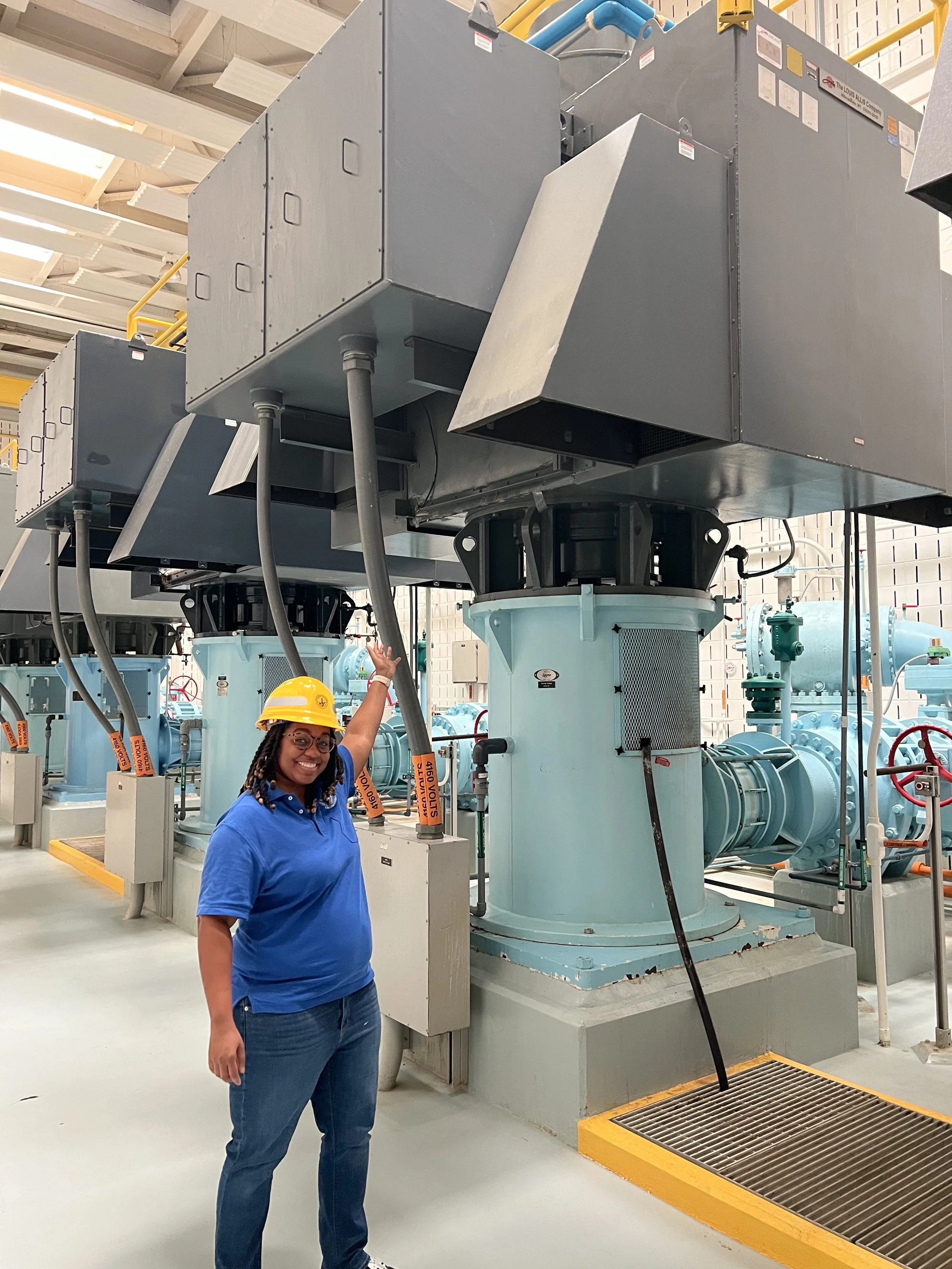 A woman wearing a blue shirt, jeans, glasses, and a yellow safety helmet smiling and pointing at large machinery in an industrial setting, with pipes and valves visible in the background.