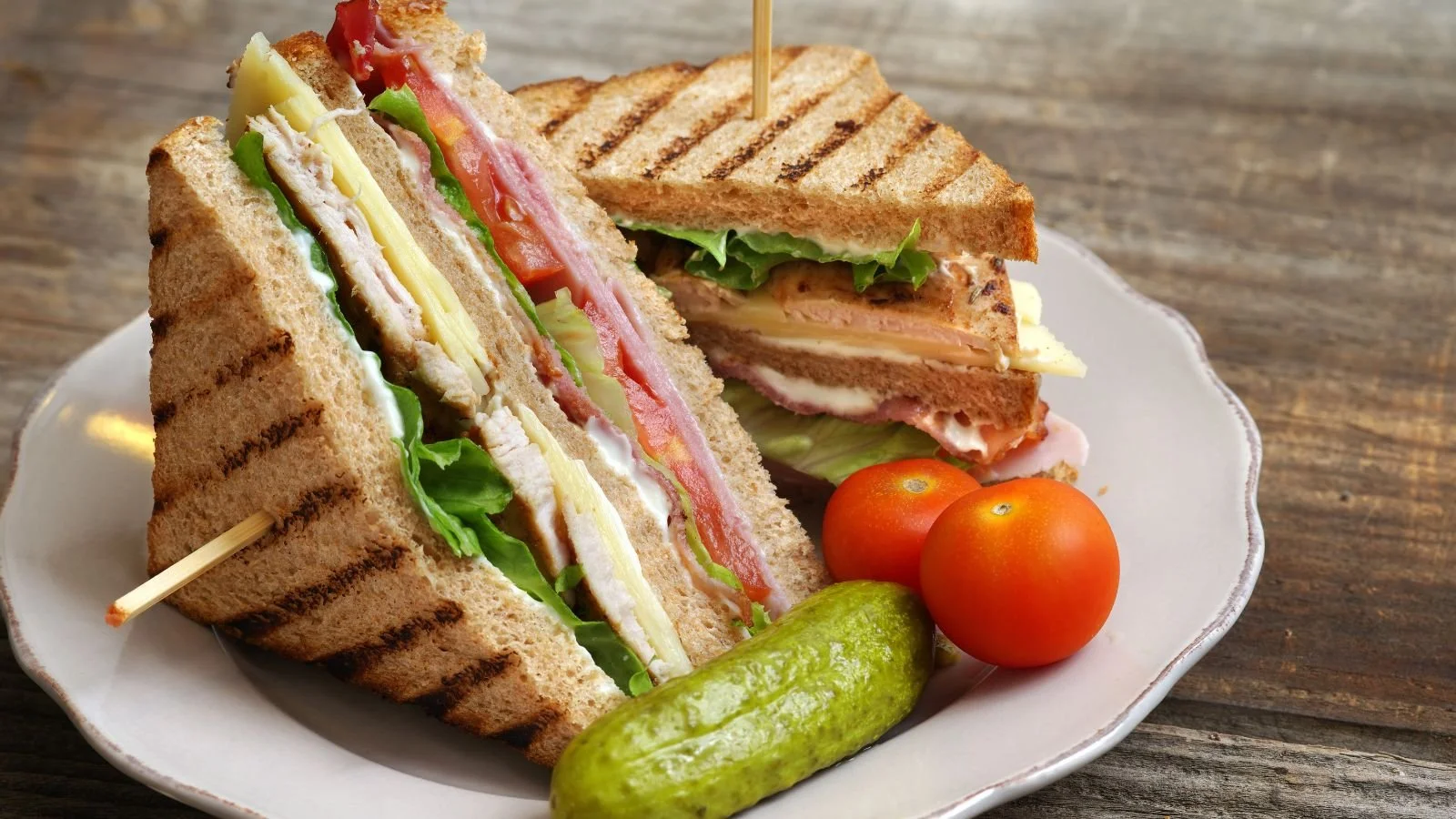 Sandwiches on a white plate with cherry tomatoes and a pickle, served on a wooden table.