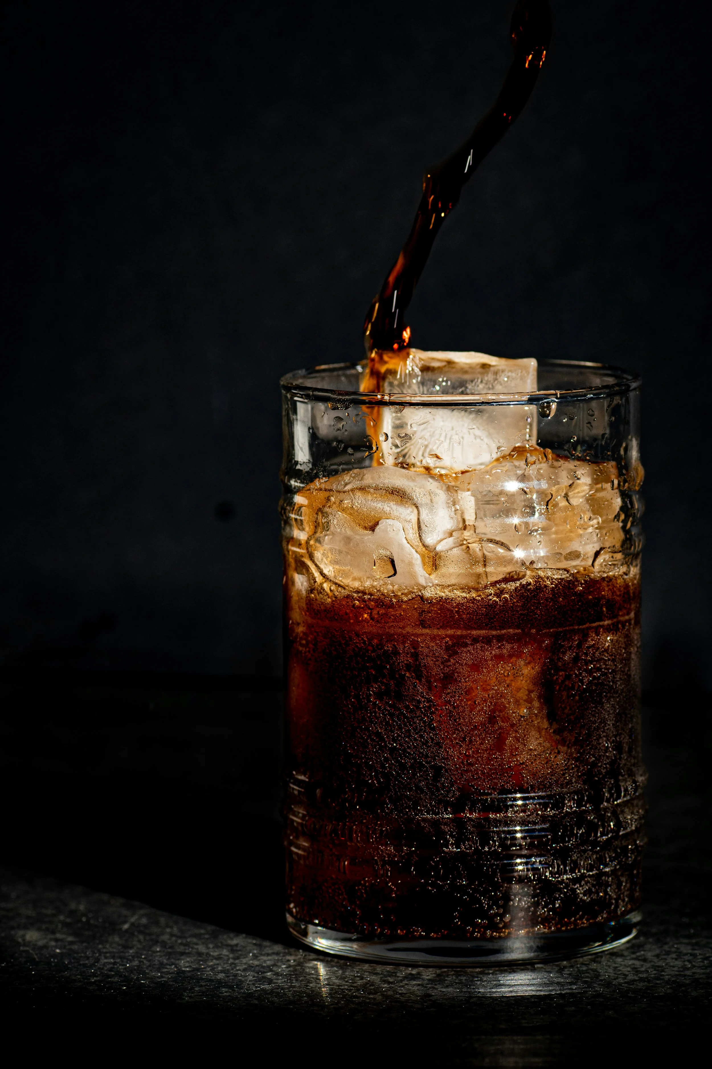 A glass of dark soda with ice cubes being poured into it against a black background.