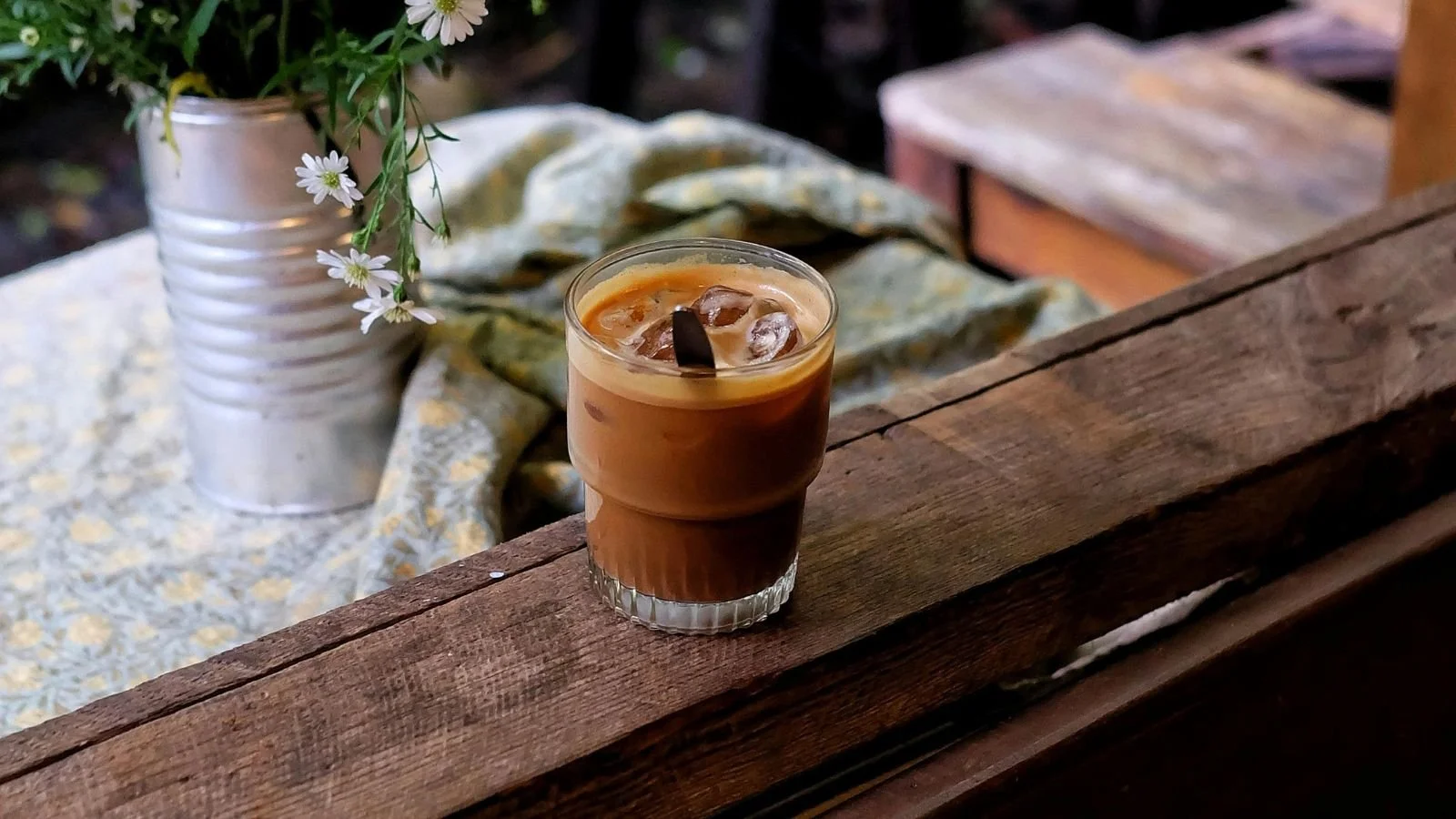 A glass of iced coffee with cream on a wooden surface, with a white flowered plant in a white ceramic pot and a soft fabric in the background.