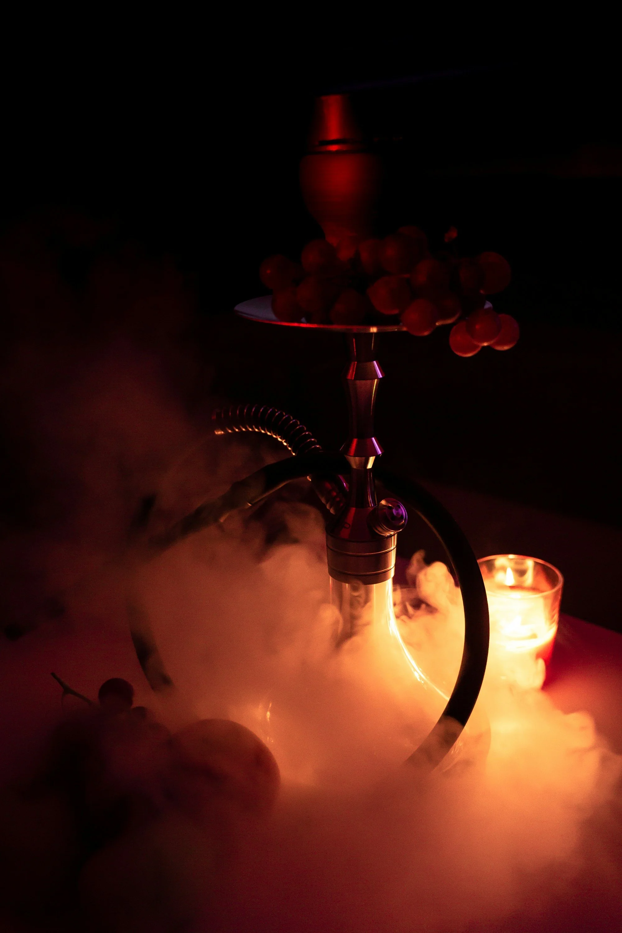 A hookah pipe with a bowl of grapes on top, emitting smoke, lit by a candle, in a dimly lit setting.