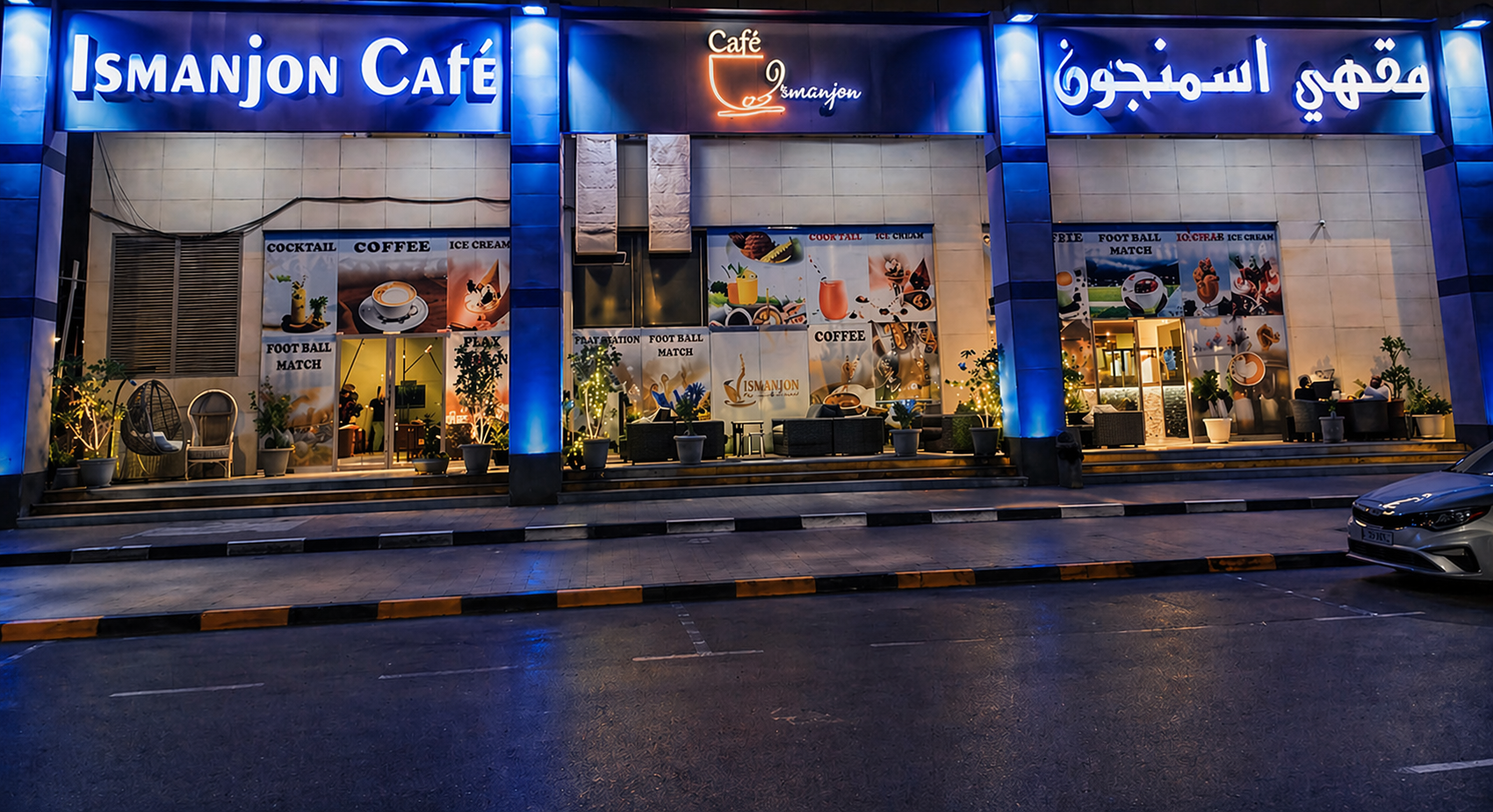 Exterior view of Ismanjon Café at night with illuminated blue and orange signboards, outdoor seating with potted plants, and advertisements for coffee, cocktails, and foot volleyball matches displayed on the windows.