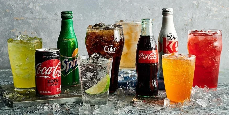Various colorful soda and soft drink bottles and glasses with ice cubes on a table.