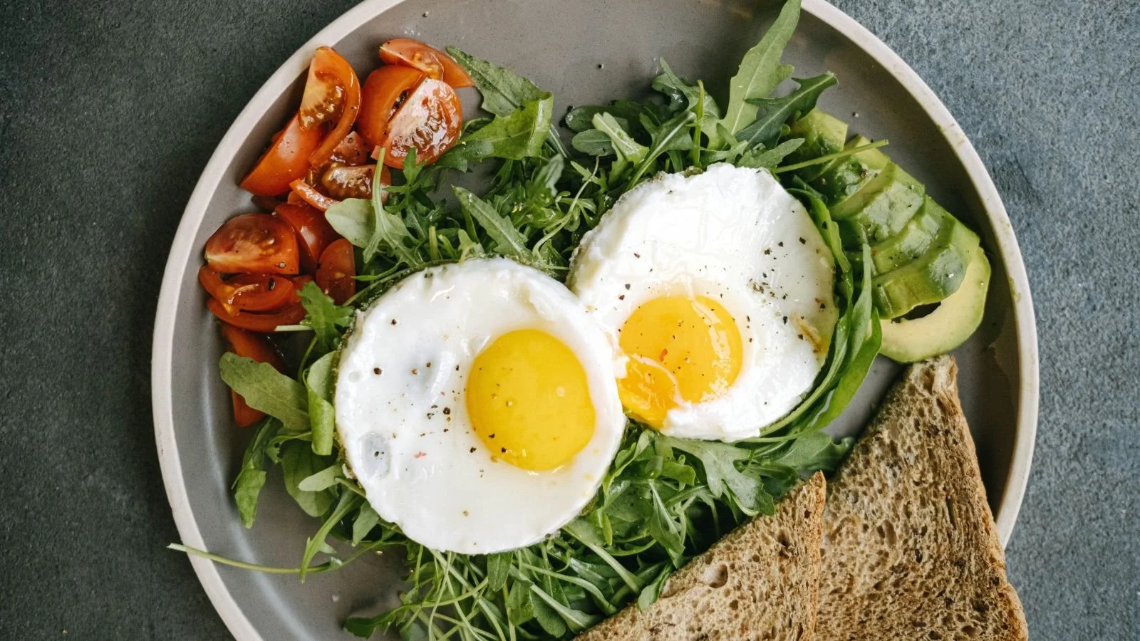 Plate with two sunny-side-up eggs, sliced avocado, mixed greens, cherry tomatoes, and toasted whole grain bread.