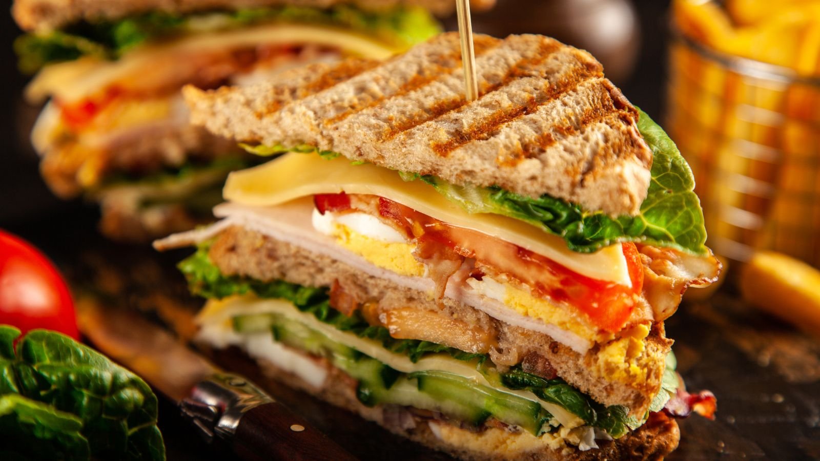 Close-up of a multi-layered sandwich with toasted bread, lettuce, tomato, cheese, bacon, and a fried egg, with grilled marks on the top slice.