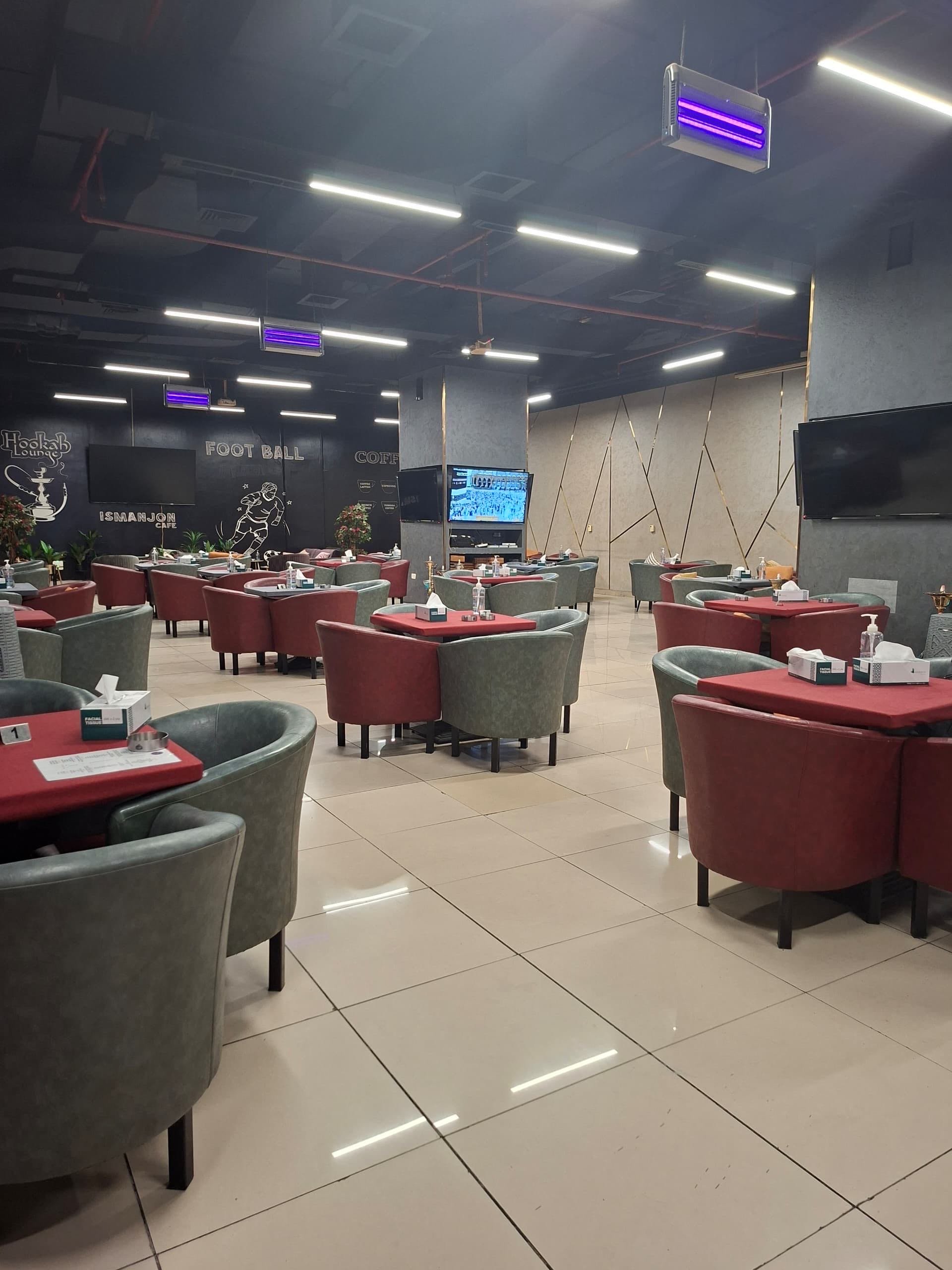Interior of a modern restaurant or cafe with red and green chairs around tables with napkins and condiments, large TV screens on walls, decorative wall art, and a tiled floor.