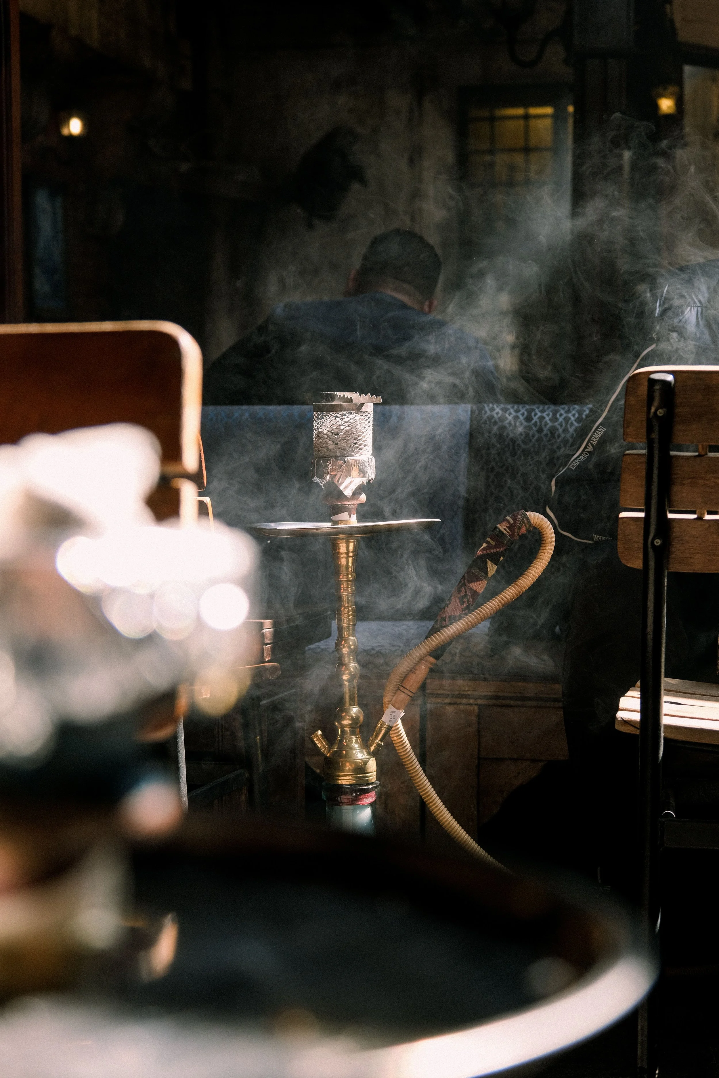 A man is sitting indoors at a table, with a hookah pipe placed in front of him. The background suggests a cozy, dimly lit room with textured walls and small lights.