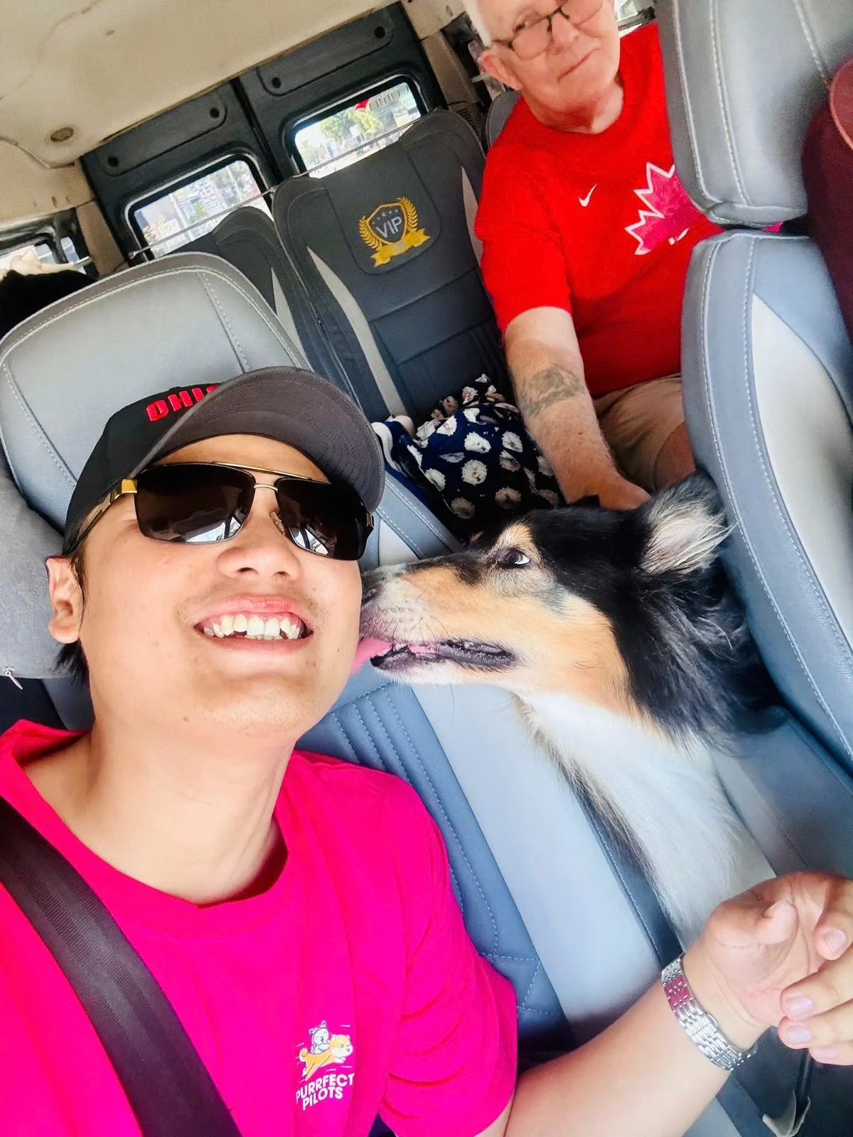 Two men and a dog inside a vehicle. The man in the foreground is wearing a pink shirt, sunglasses, a wristwatch, and a cap, smiling at the camera. The dog, which appears to be a Border Collie mix, is licking the man's face. Behind them, an older man in a red shirt and glasses is seated, looking toward the camera.