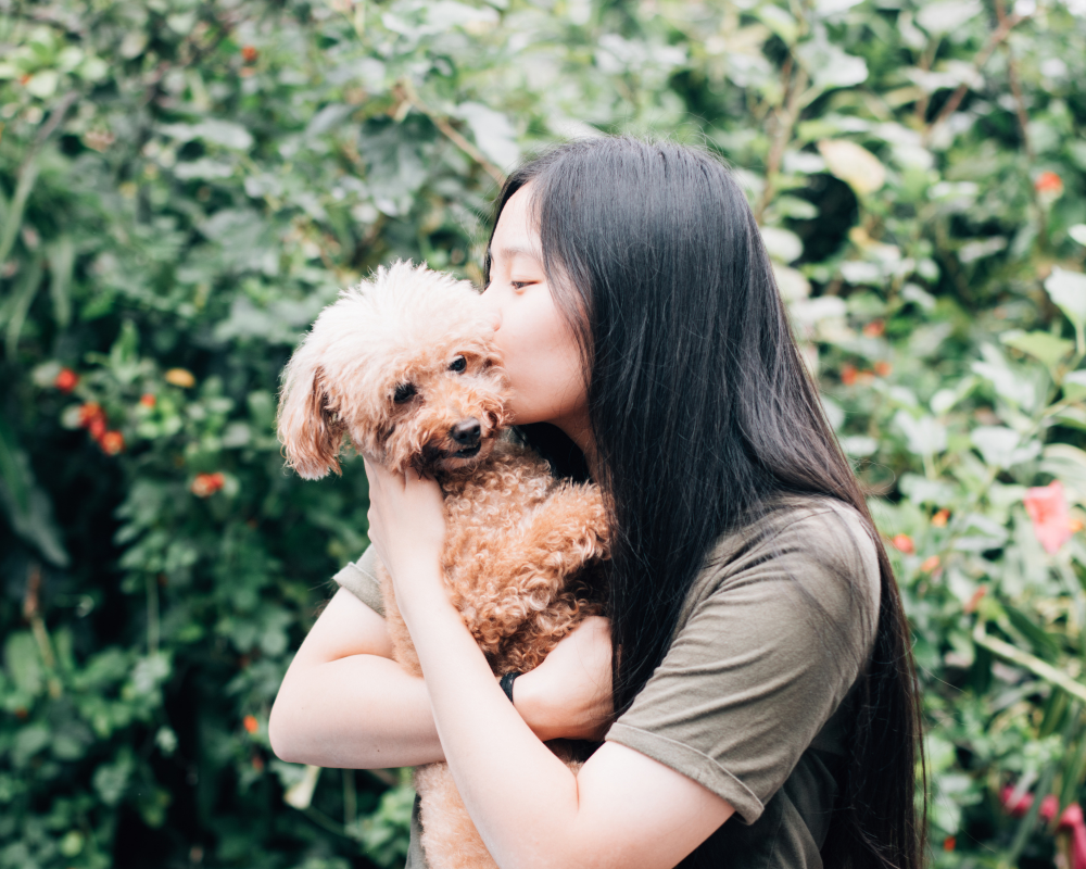 A young woman with long black hair holding a small, curly-haired dog and giving it a kiss on the head in an outdoor garden setting.