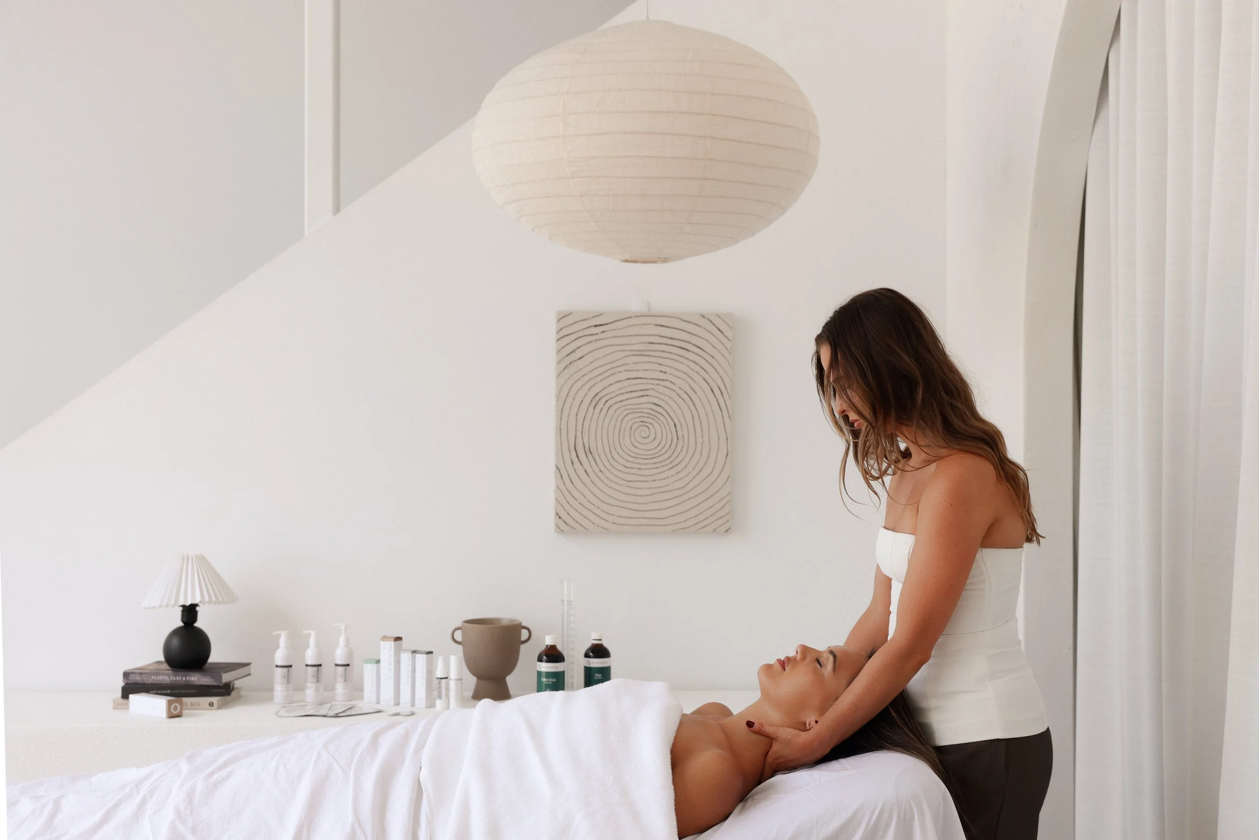 A woman receiving a facial treatment from a therapist in a bright, minimalist room with white walls, books, and skincare products on a table.