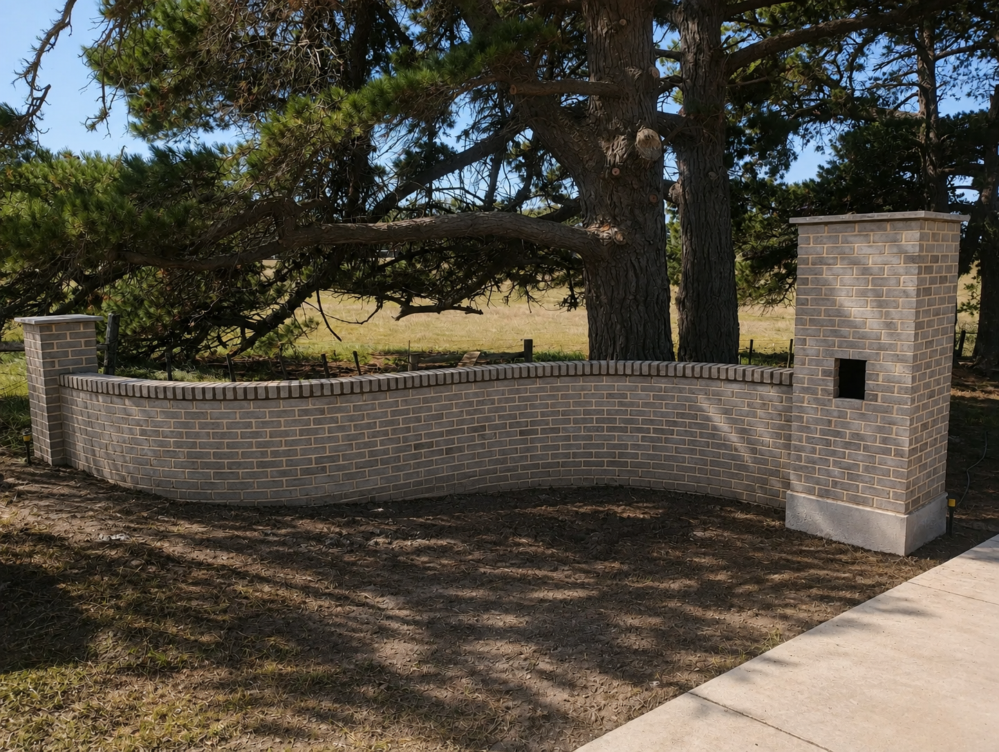 A curved brick wall with two brick columns, one on each end, and a small square opening in the right column, situated in a natural setting with trees and grass in the background.