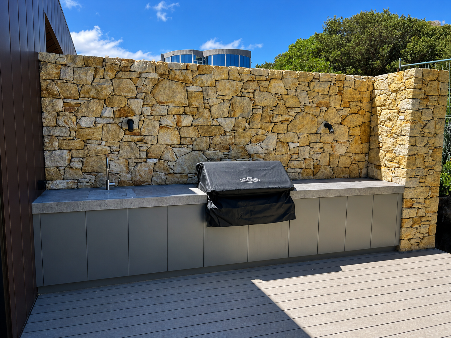 Outdoor kitchen with stone wall, gray cabinets, and a covered barbecue grill on a wooden deck, under blue sky with scattered clouds.