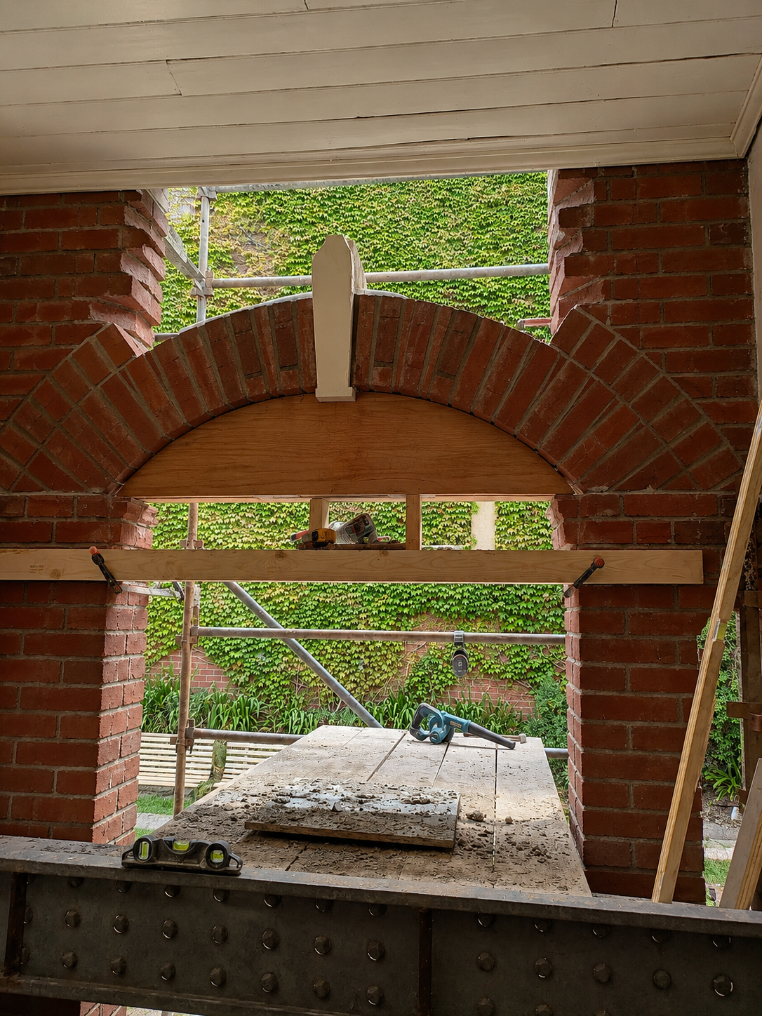 Construction site showing heritage brick arches, wooden beams, power tools, and scaffolding, with leafy greenery visible outside.